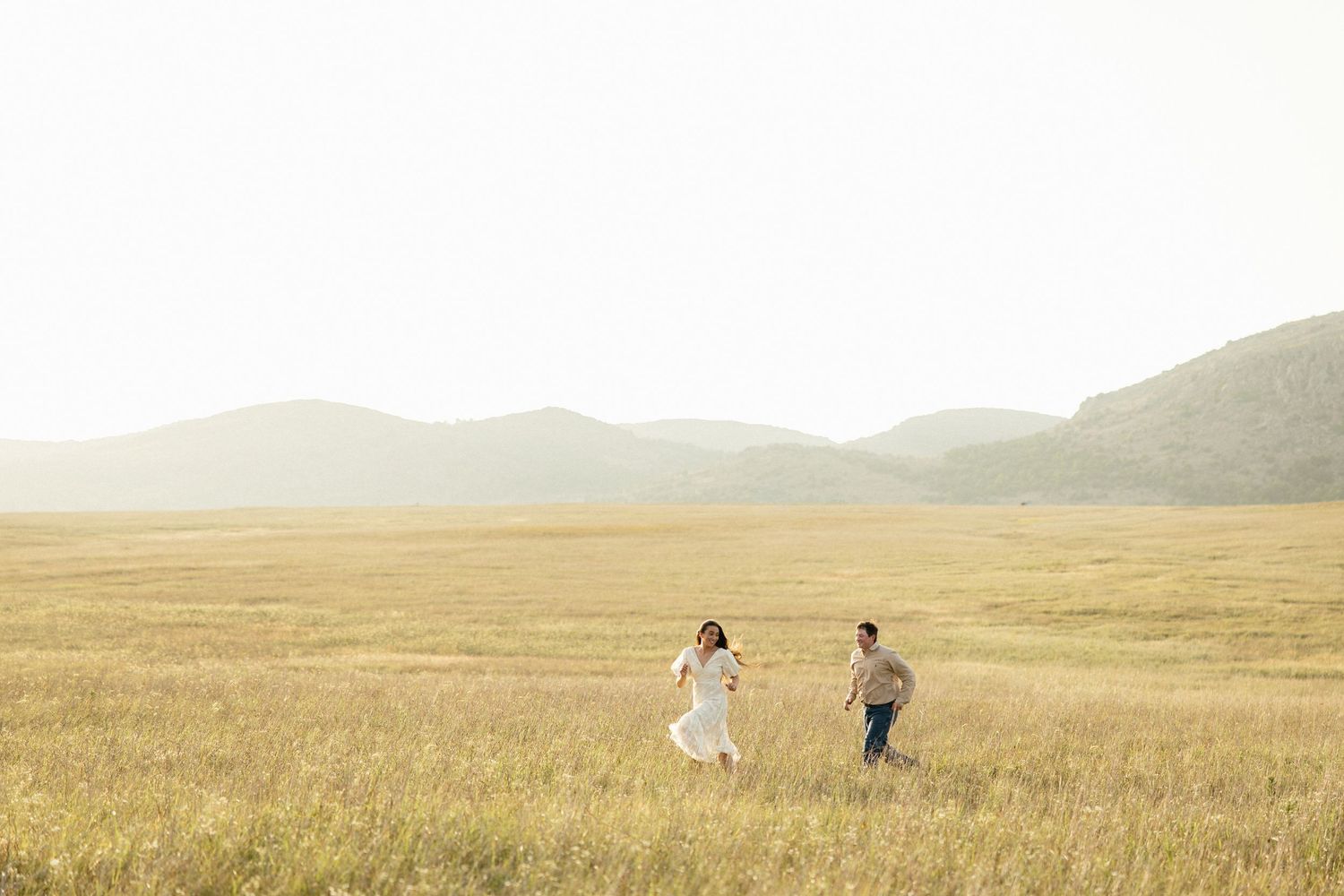 Couple holding hands running through vast golden field with rolling mountains in the background.