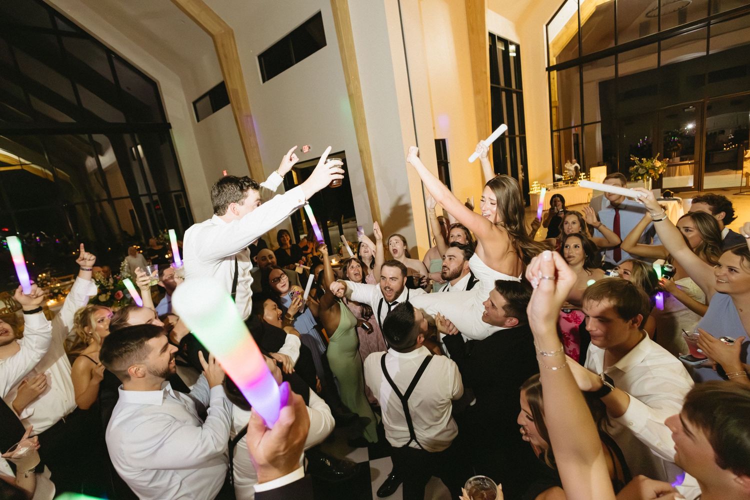 Bride lifted above crowd of celebrating wedding guests holding colorful glow sticks at night reception.