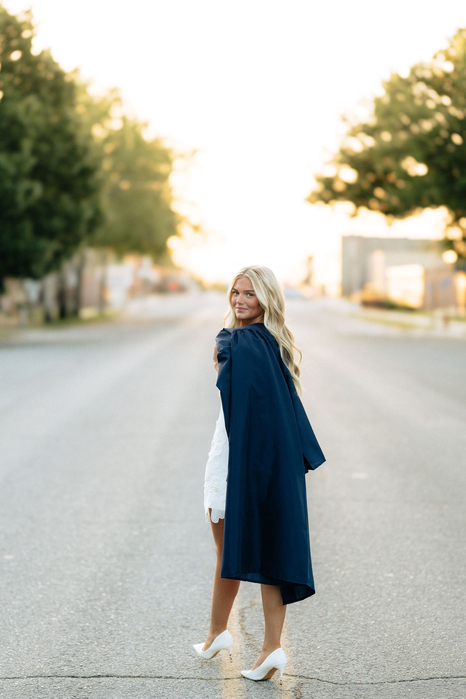 Blonde woman in navy graduation gown and white heels smiling on a sunlit street at golden hour.