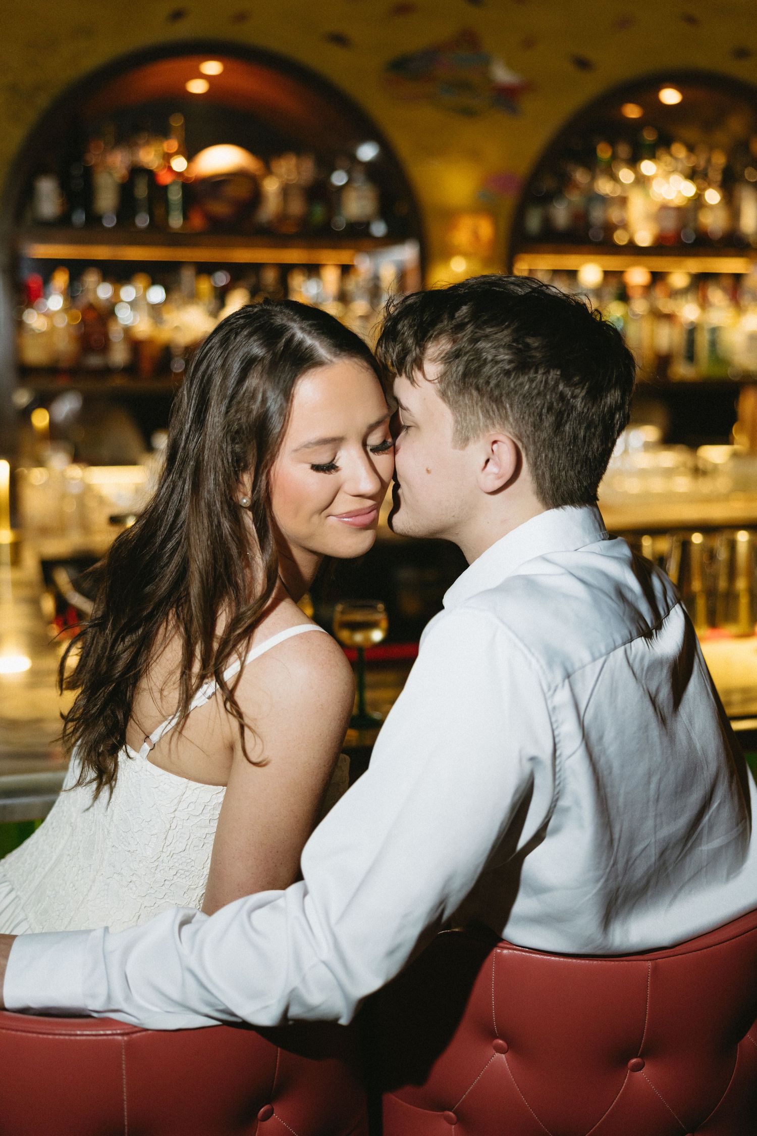 A couple shares a romantic moment at a dimly lit bar, with the man leaning in to kiss the smiling woman in white.