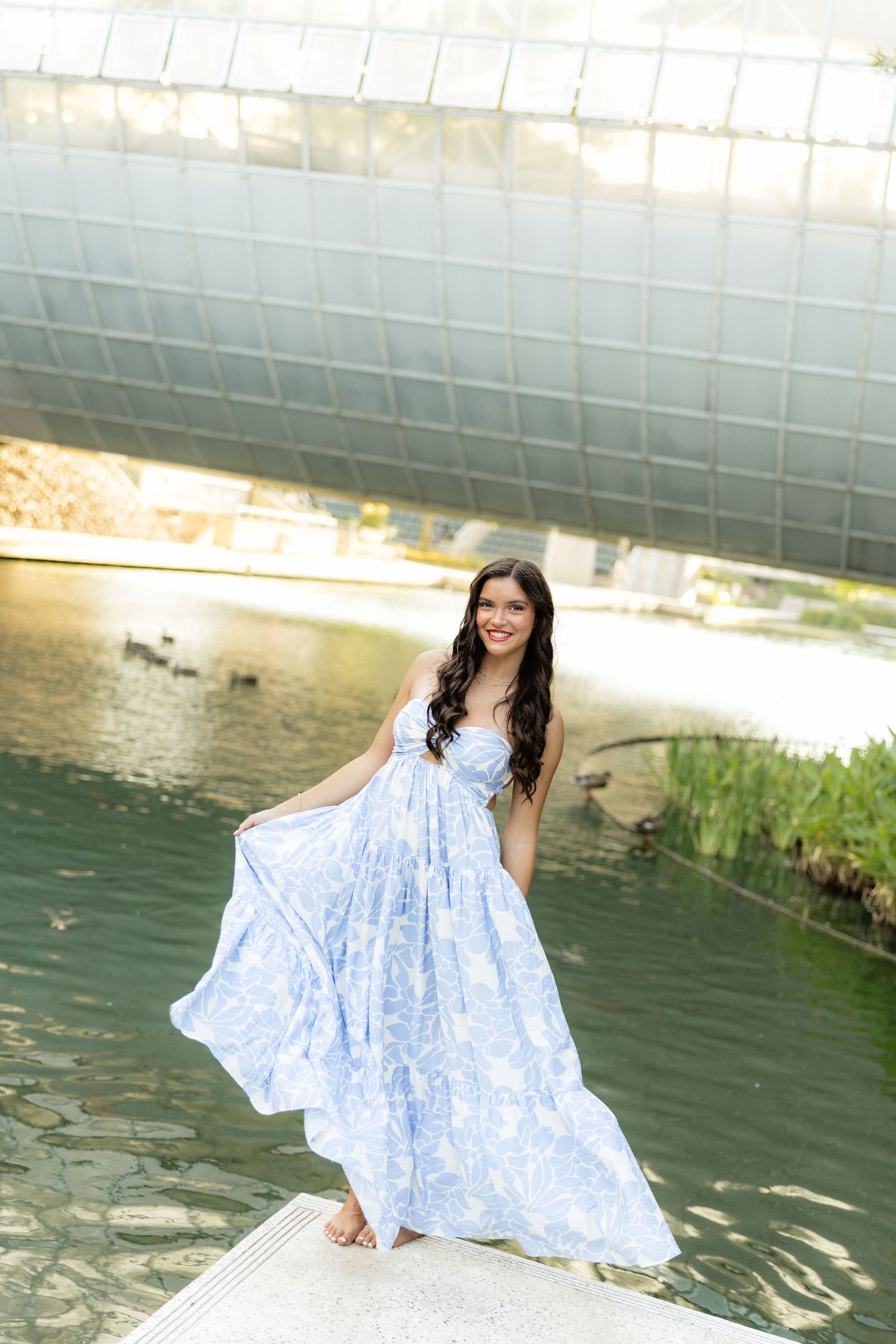 Young woman in flowing blue floral maxi dress posing barefoot on ledge by green pond with ducks.