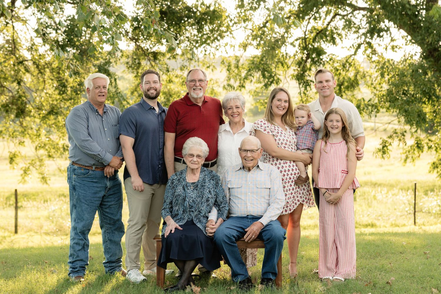 Multi-generational family portrait outdoors under a large tree on green grass, featuring grandparents, parents, and children.