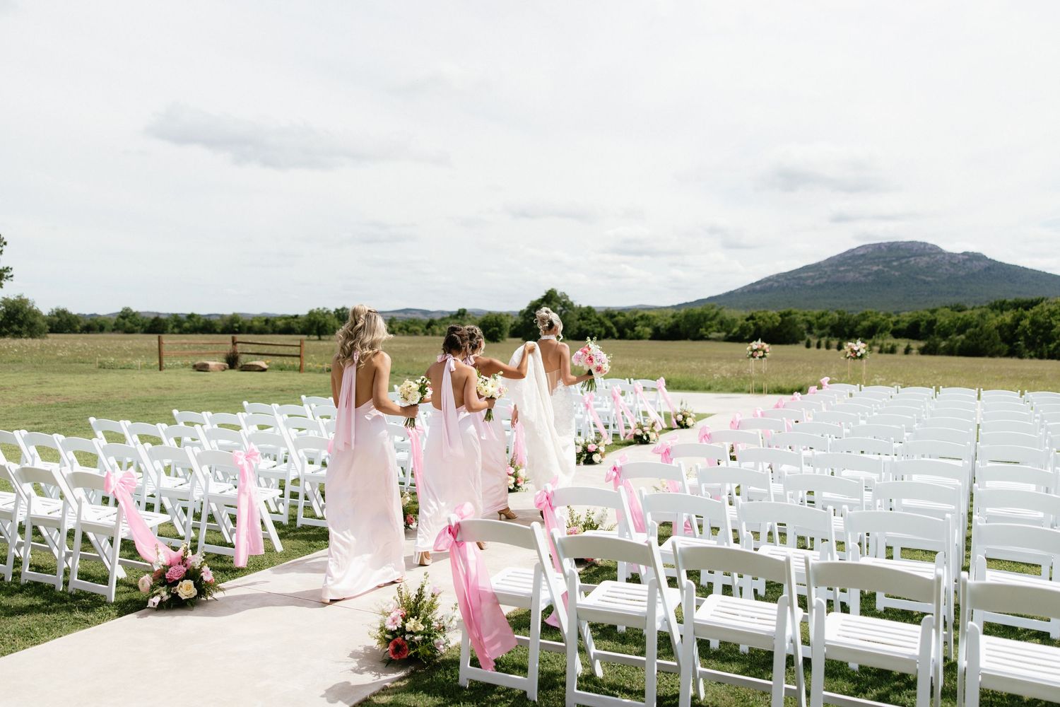Bridesmaids in pink dresses walking down outdoor wedding aisle with mountain view.