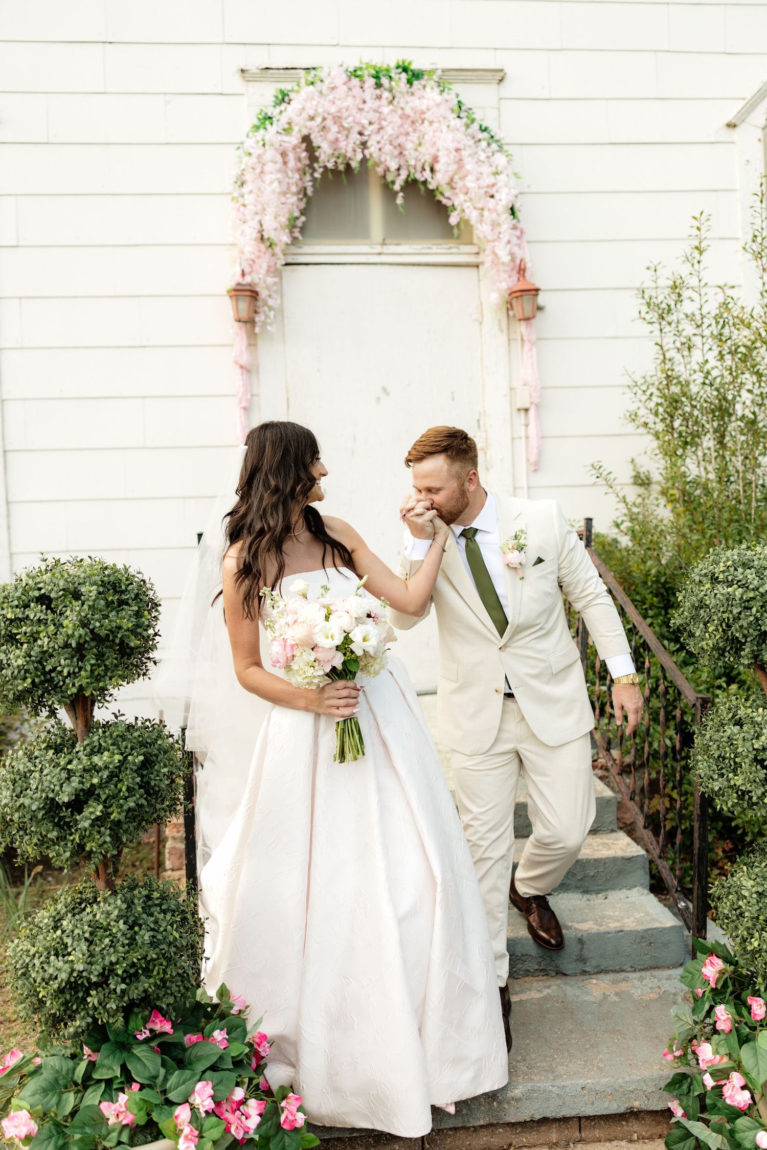 Groom kisses bride's hand on steps of white chapel adorned with pink floral arch, bride holding white bouquet.