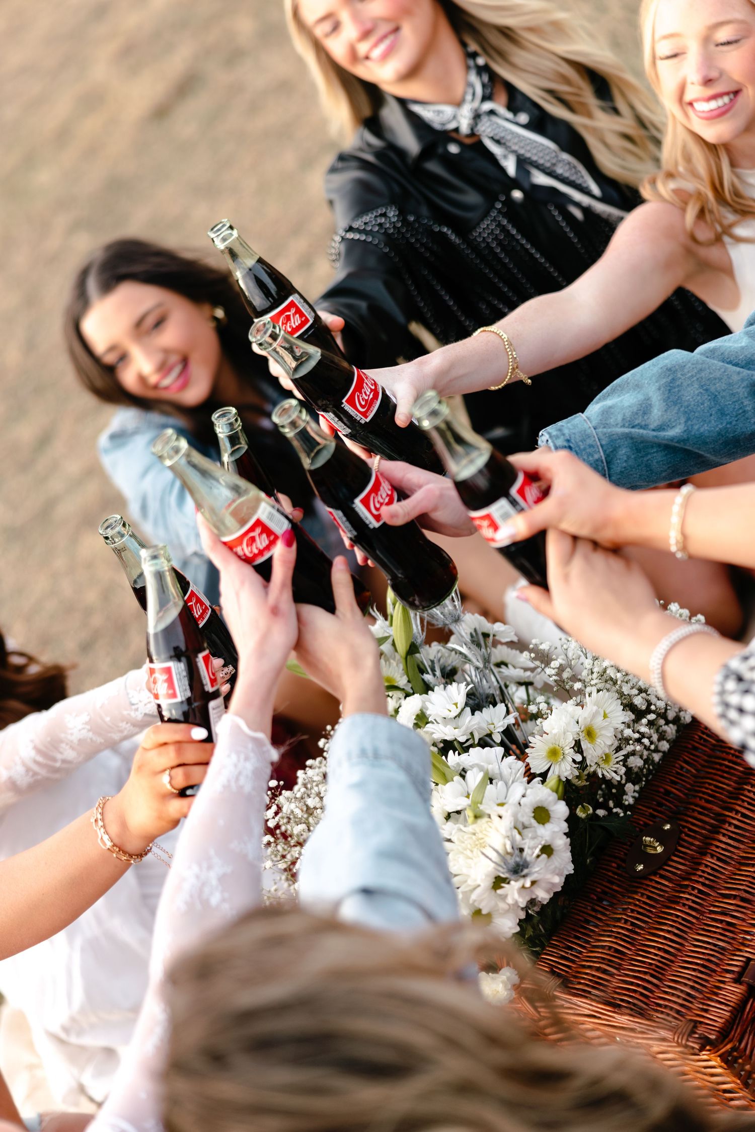 Group of smiling women clinking Coca-Cola glass bottles together at outdoor western-themed party.