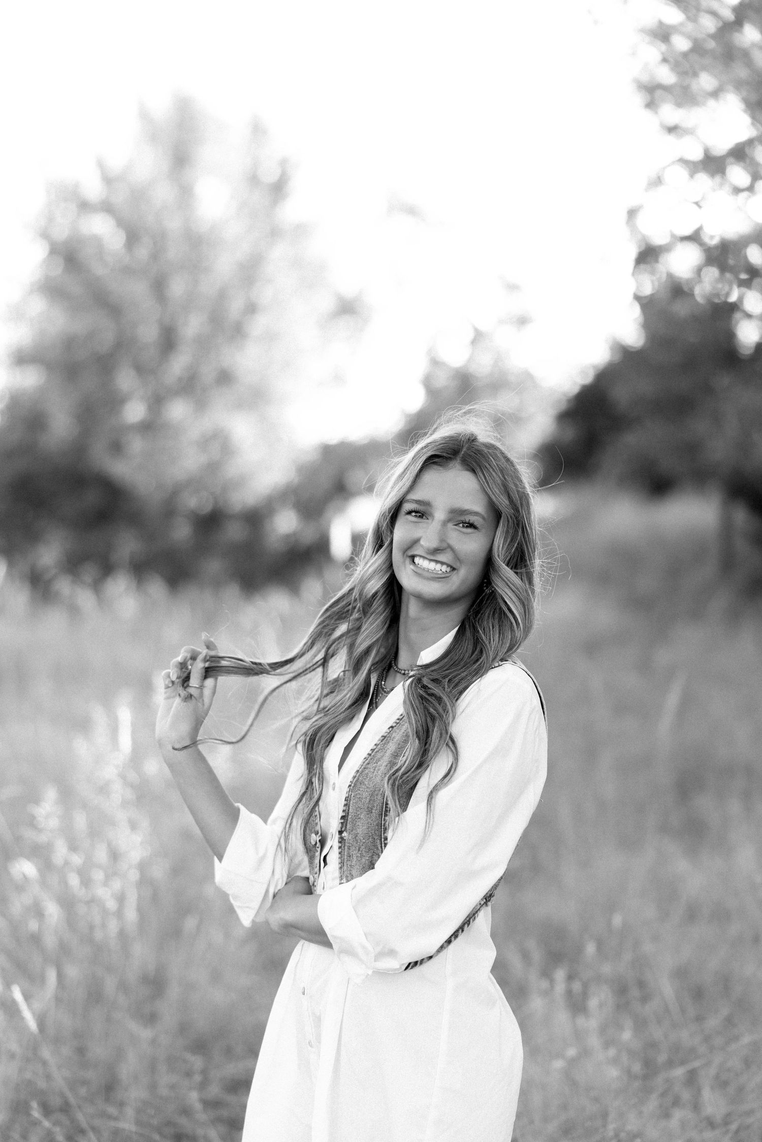 Smiling young woman in white dress twirling her hair in a sunlit field, black and white portrait.