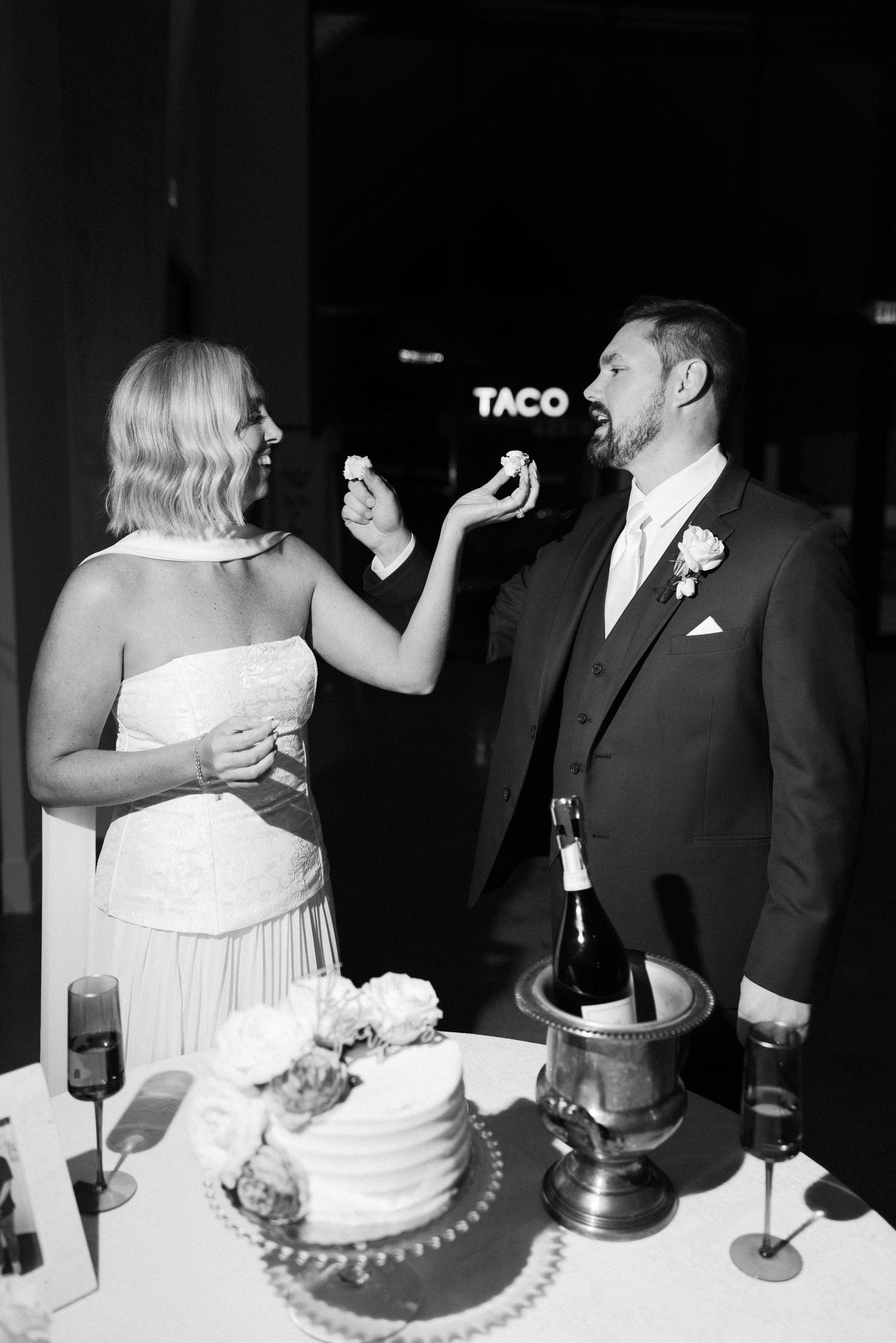 Black and white photo of bride feeding groom wedding cake at elegant reception with champagne.
