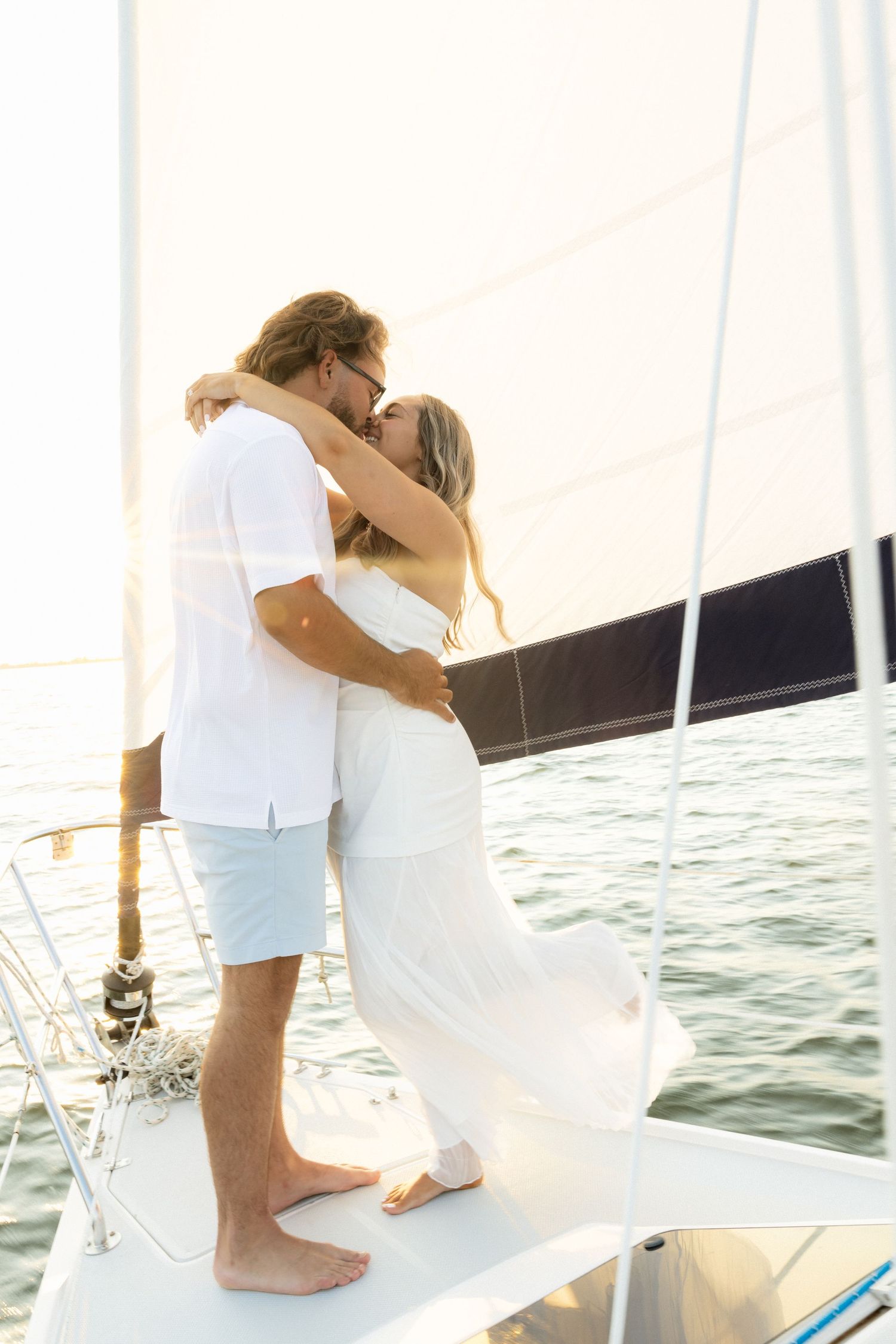 Romantic couple kissing on sailboat bow, dressed in white, with ocean backdrop on sunny day.