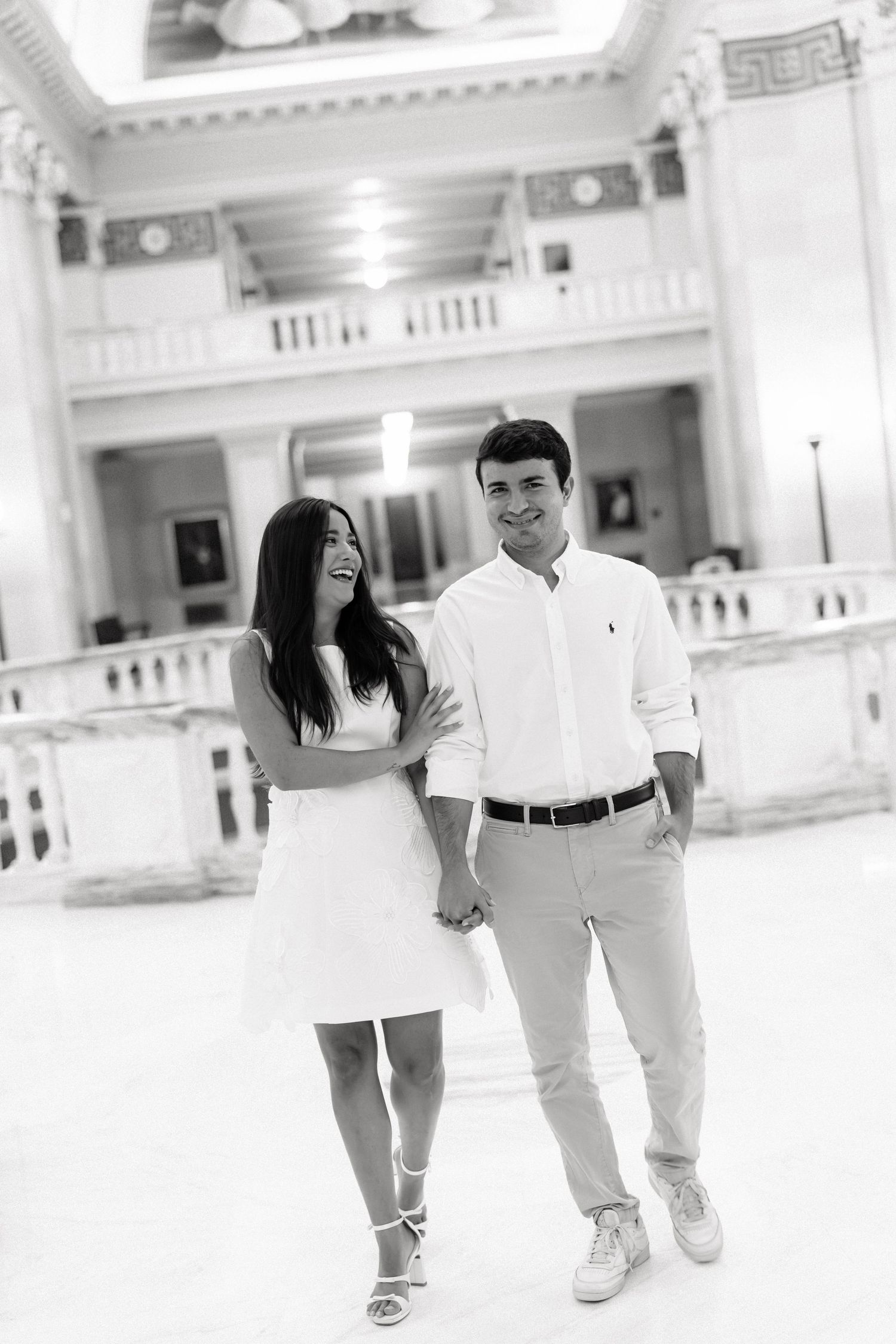 Black and white photo of a smiling couple holding hands in an elegant white interior building.