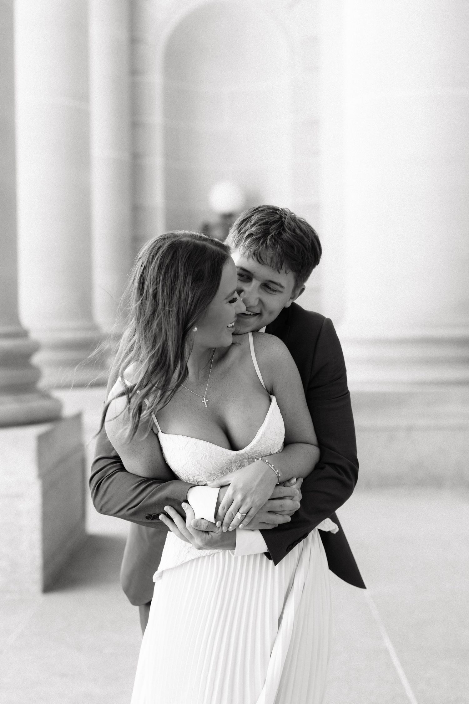 Black and white photo of a couple embracing and kissing in wedding attire near elegant stone columns.