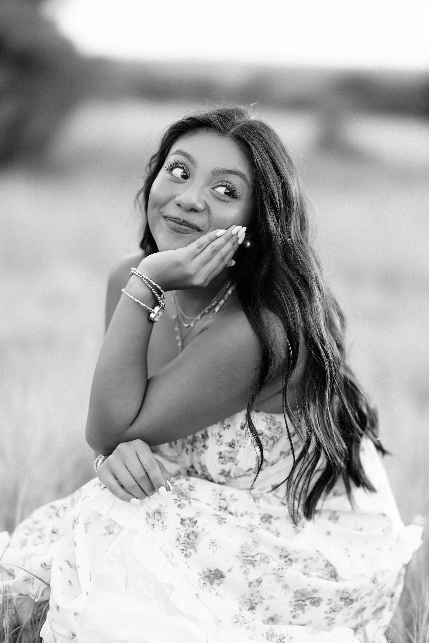 Black and white portrait of a young woman in a floral dress, posing playfully in a field.