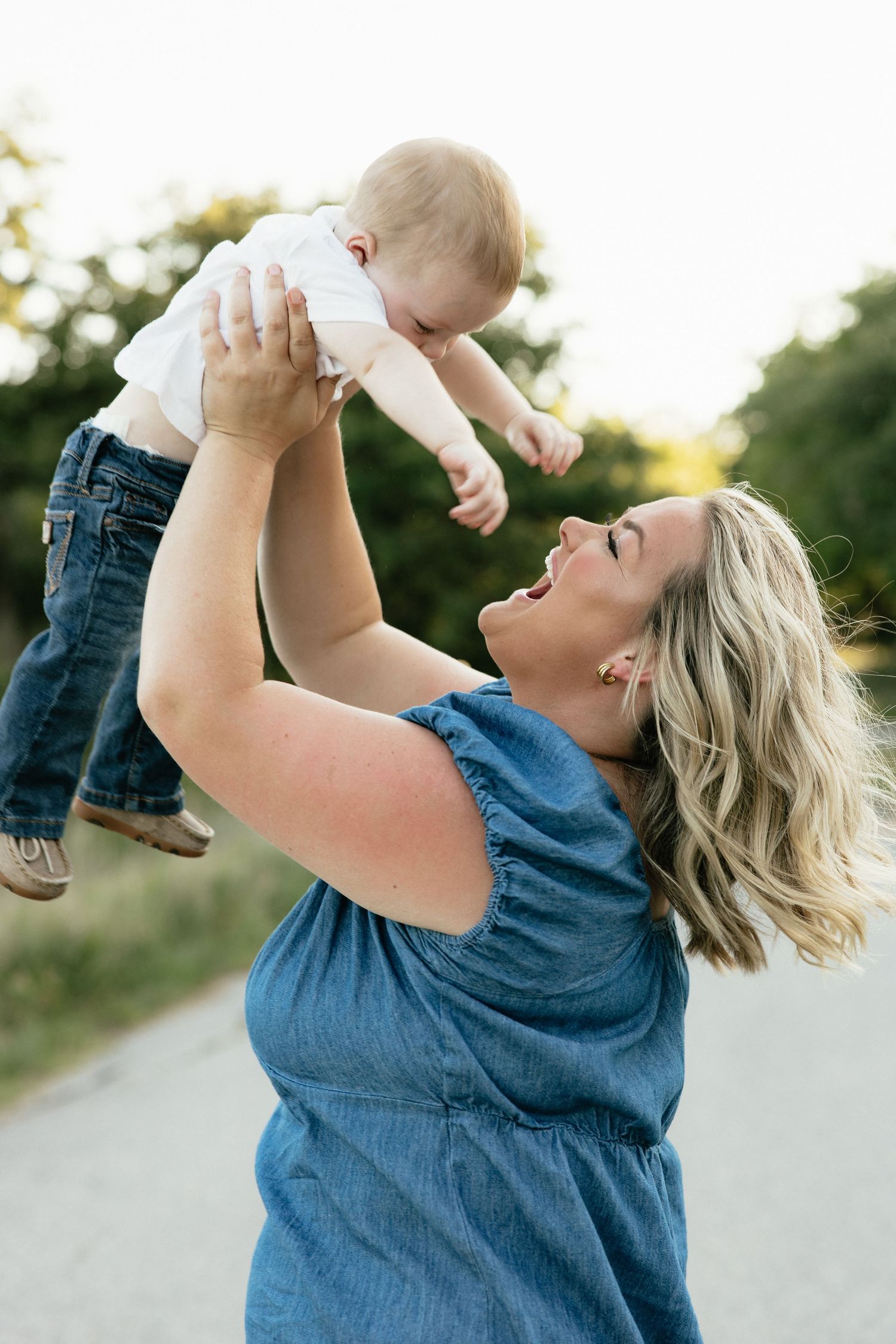 A joyful mother lifts her smiling baby overhead outdoors on a sunny path, both wearing casual blue outfits.