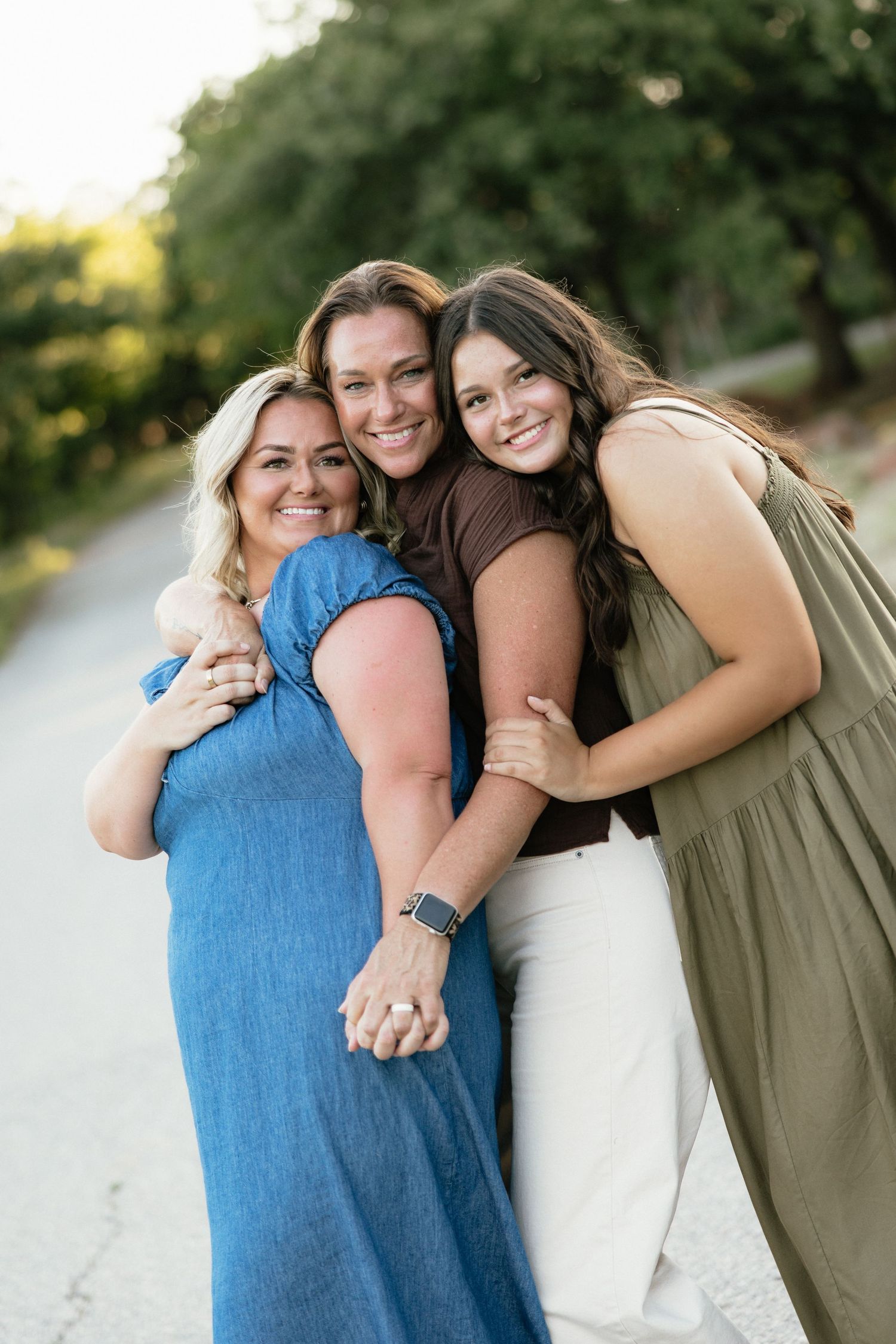 Three smiling women hugging outdoors on a sunny path surrounded by green trees.