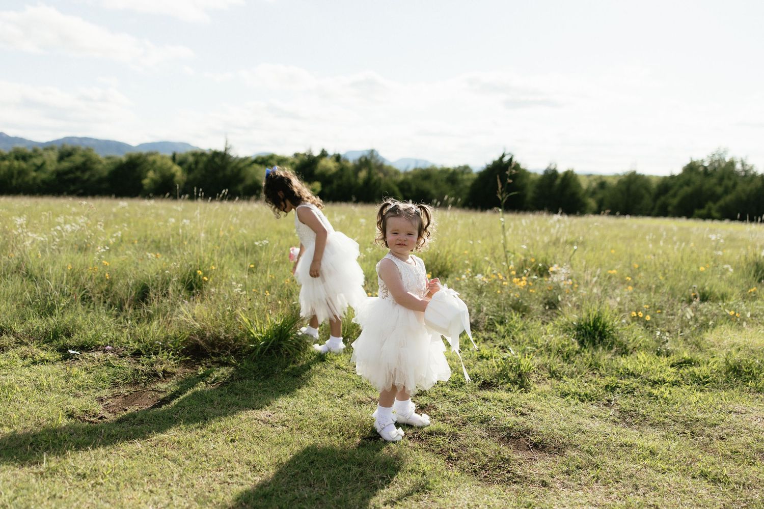 Two young flower girls in white tulle dresses running through a green meadow with mountains in the background.