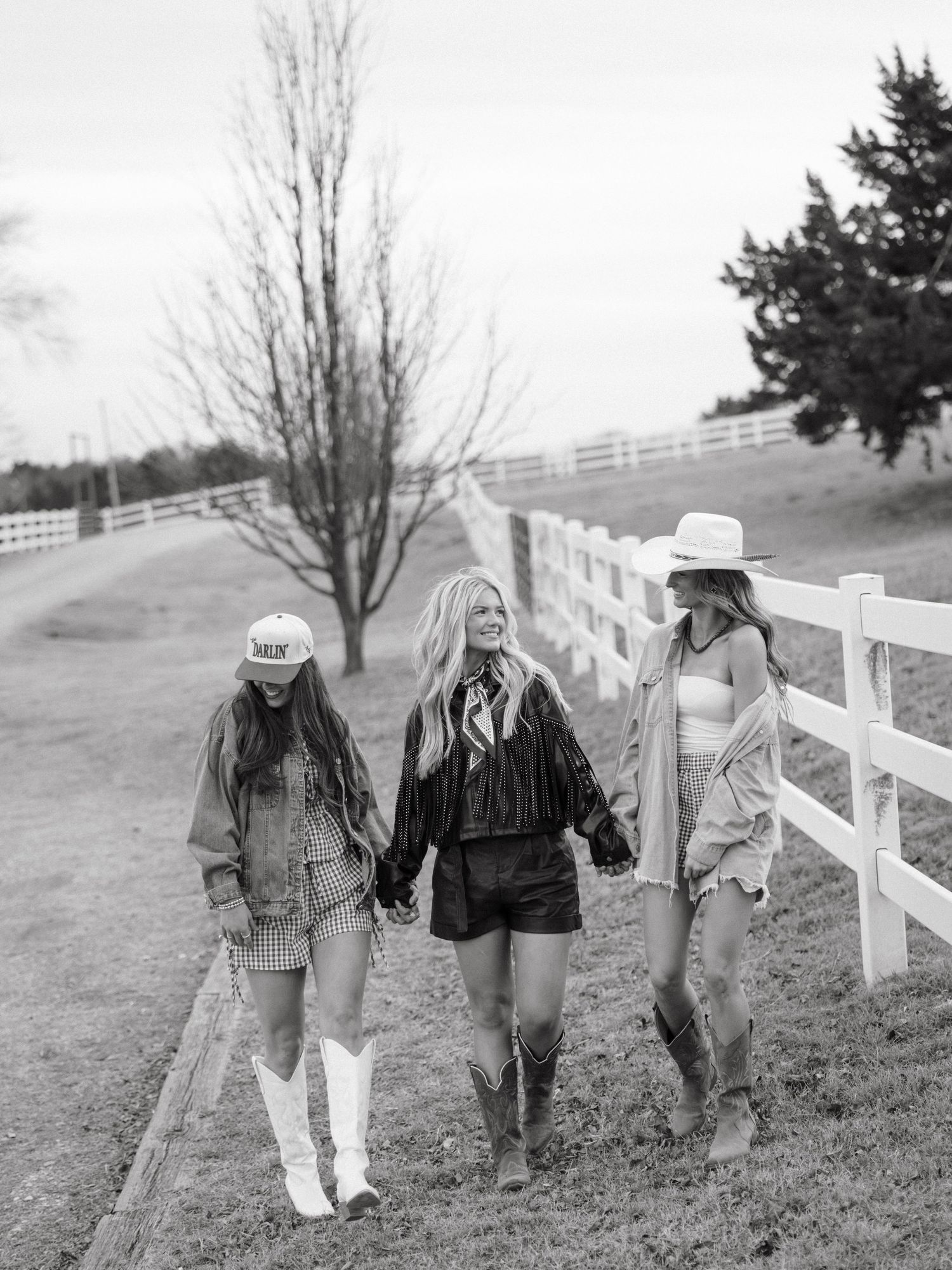 Black and white photo of three women in western outfits walking along a white fence on a farm.