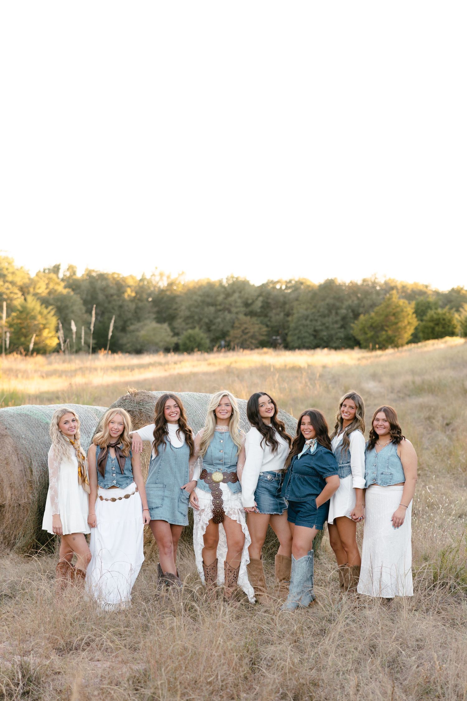 Group of young women in boho western outfits posing by a hay bale in a golden field at sunset.
