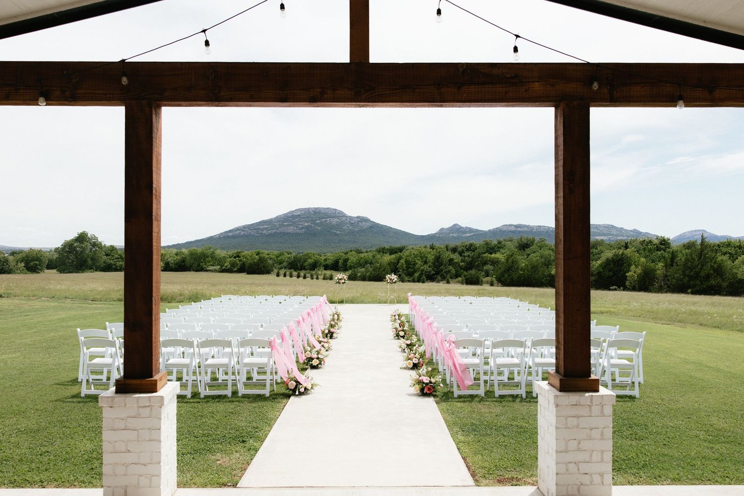 Elegant outdoor wedding ceremony setup with white chairs, floral aisle, and mountain views under a rustic wooden pavilion.