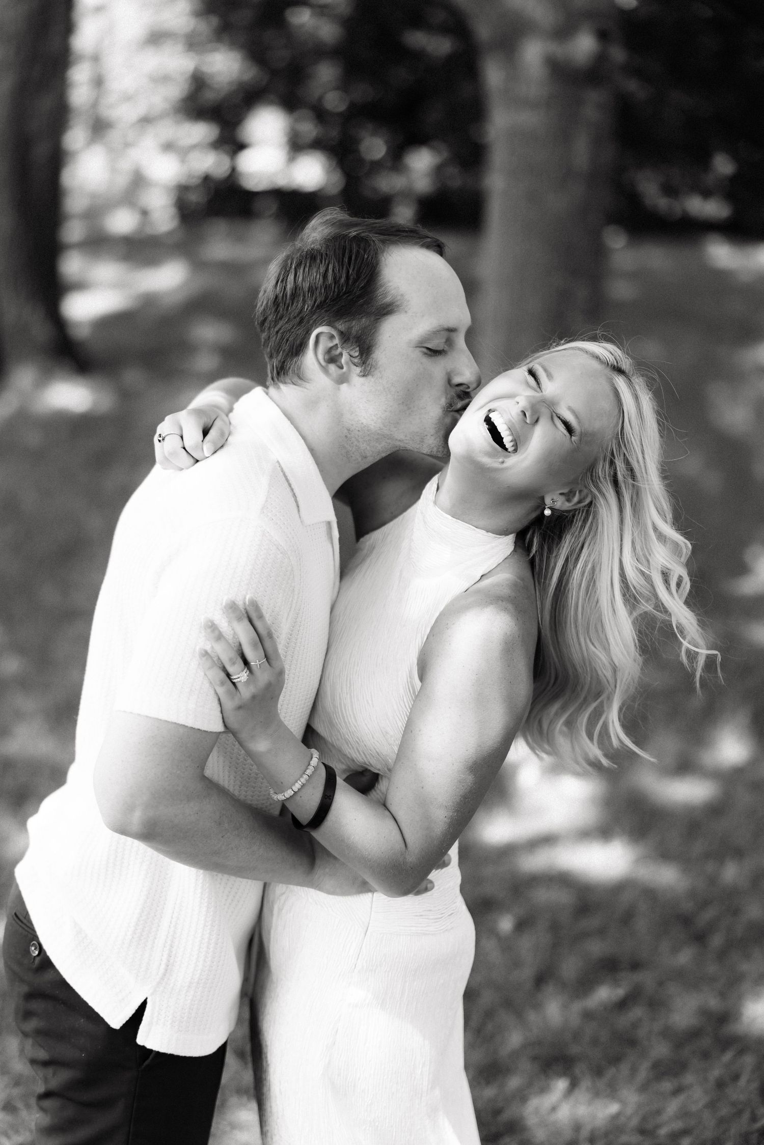 Black and white photo of a couple embracing outdoors, man kissing womans cheek as she laughs joyfully.