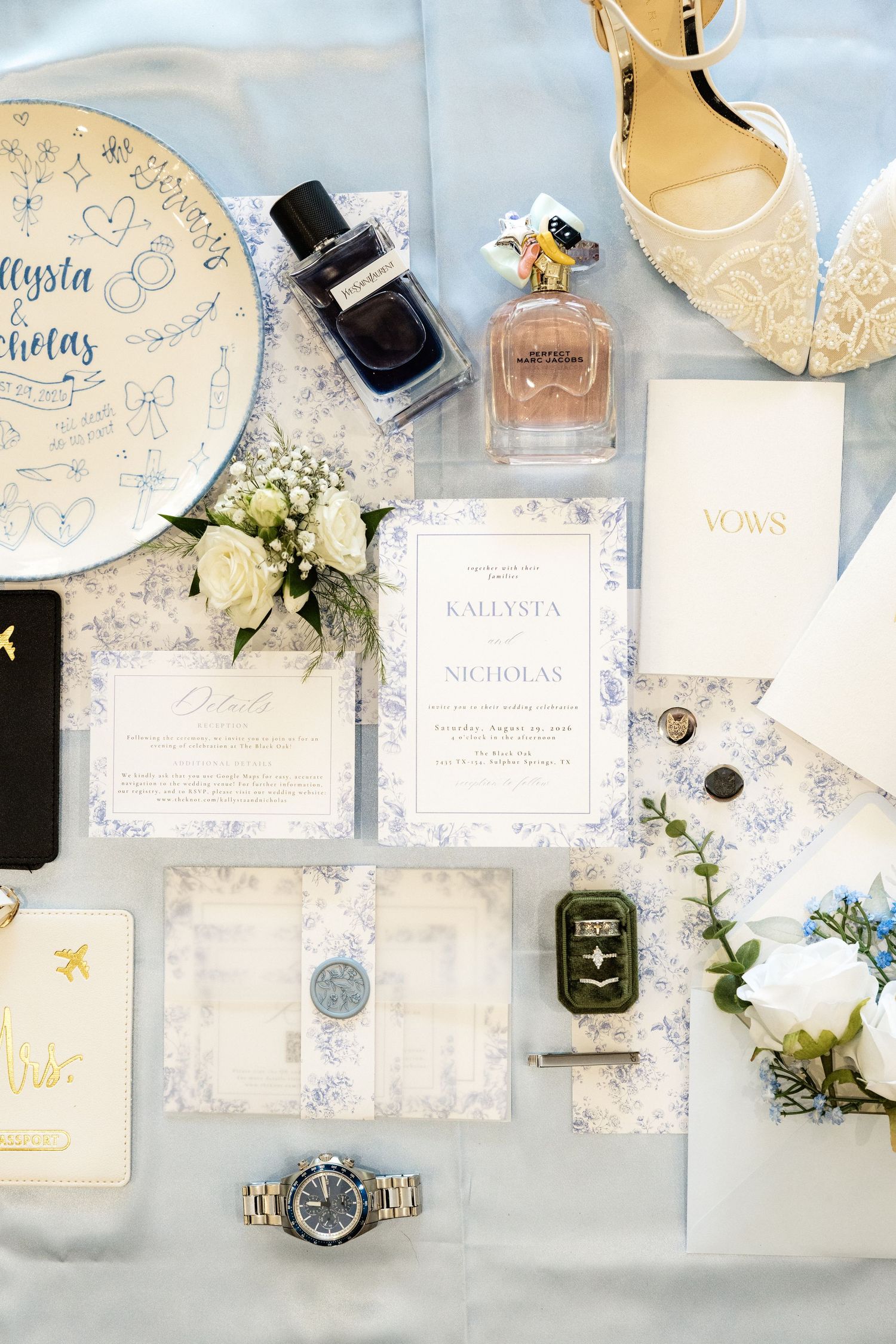 Elegant wedding flat lay with blue floral invitations, perfume, vows book, watch, and white roses on soft blue fabric.