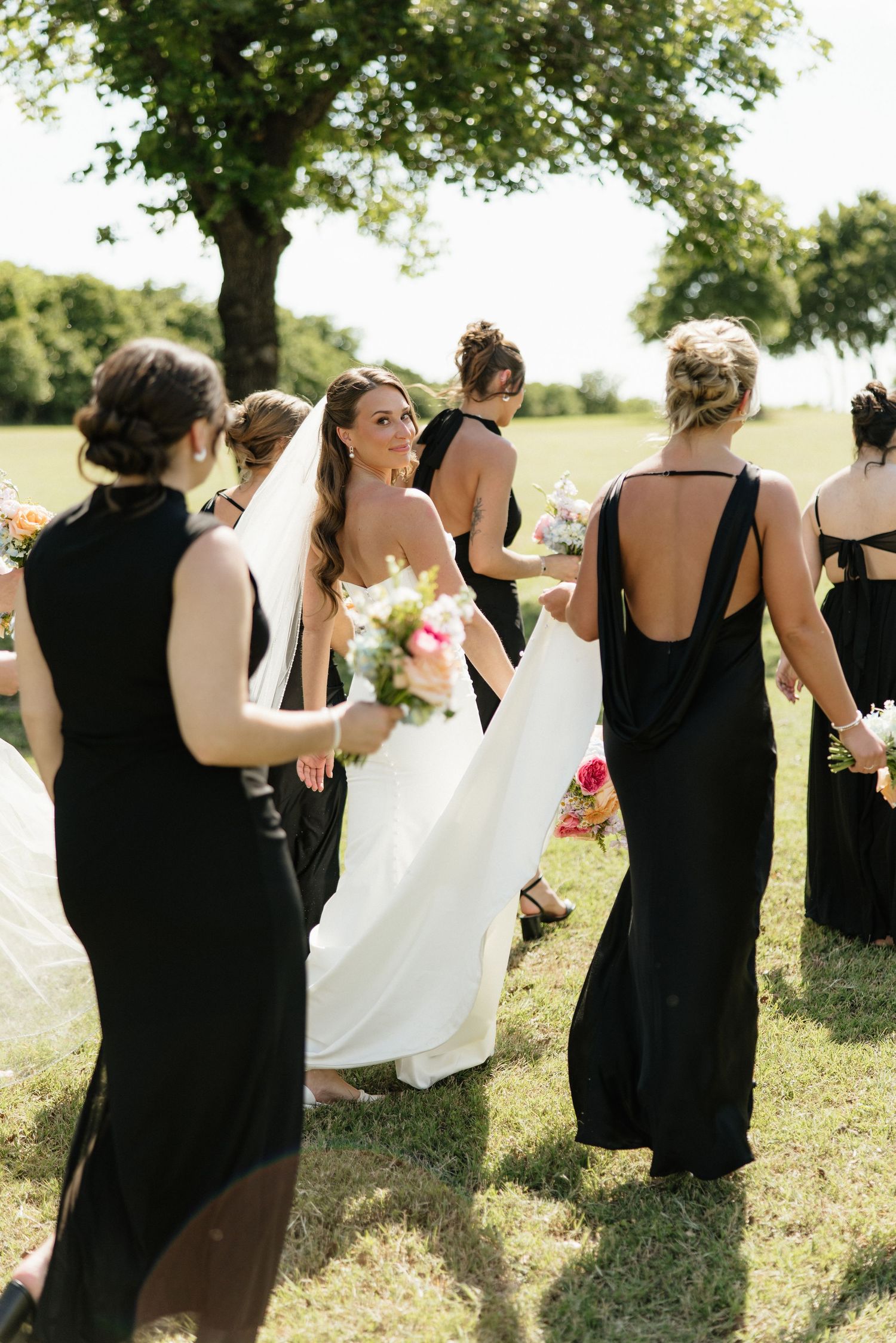 Bride in white gown walking with bridesmaids in black dresses across green lawn.