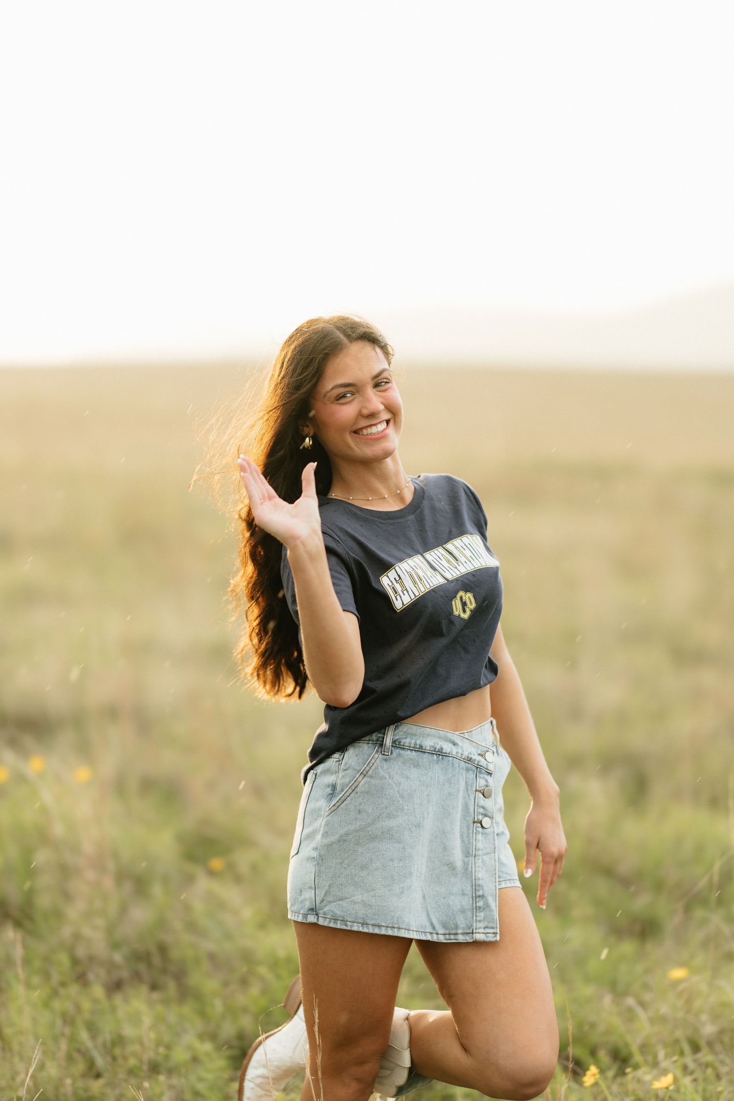 Young woman in denim skirt and crop top smiling in golden field at sunset.