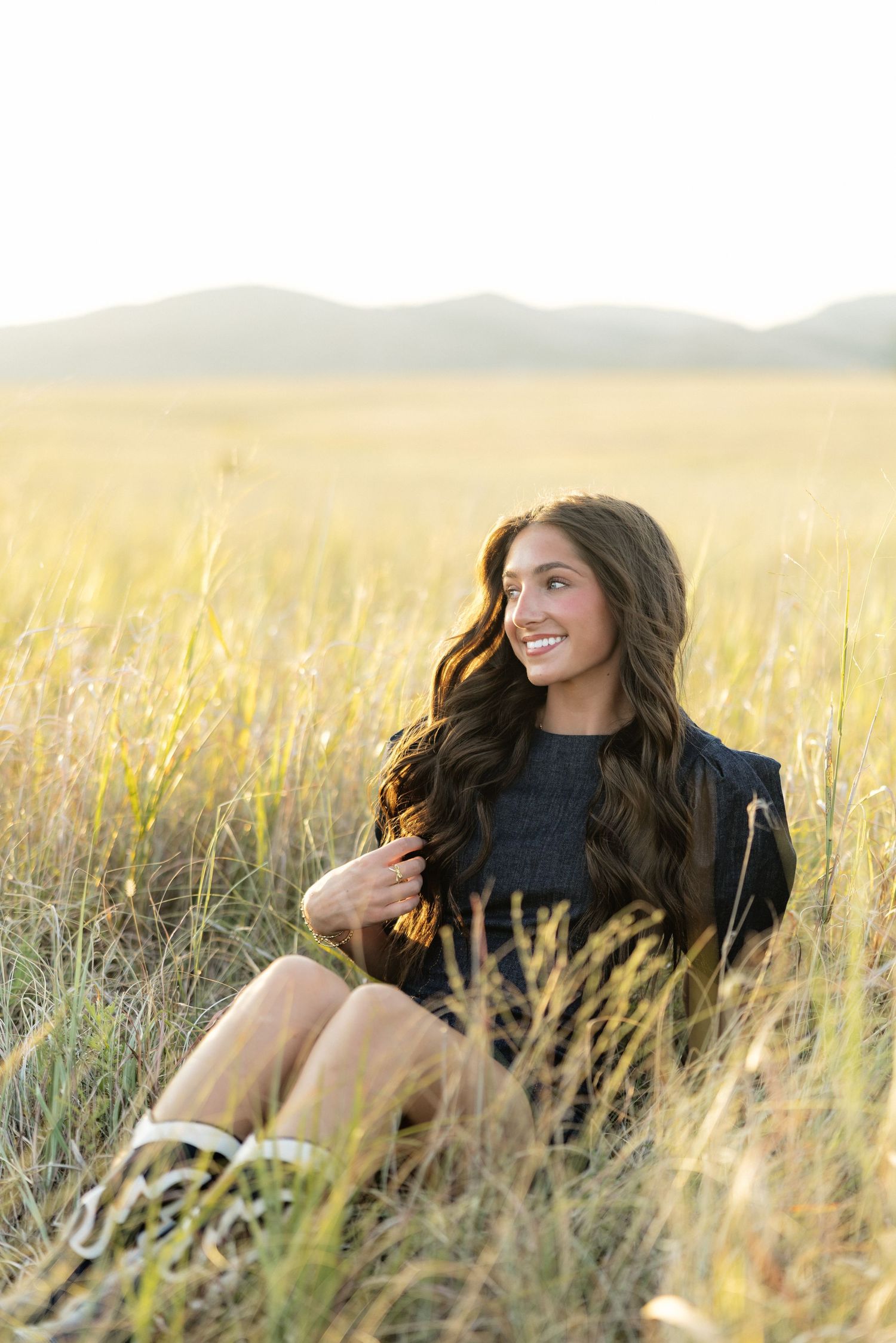 Young woman with long wavy hair sitting in golden field grass, smiling, wearing black dress, mountains in background.