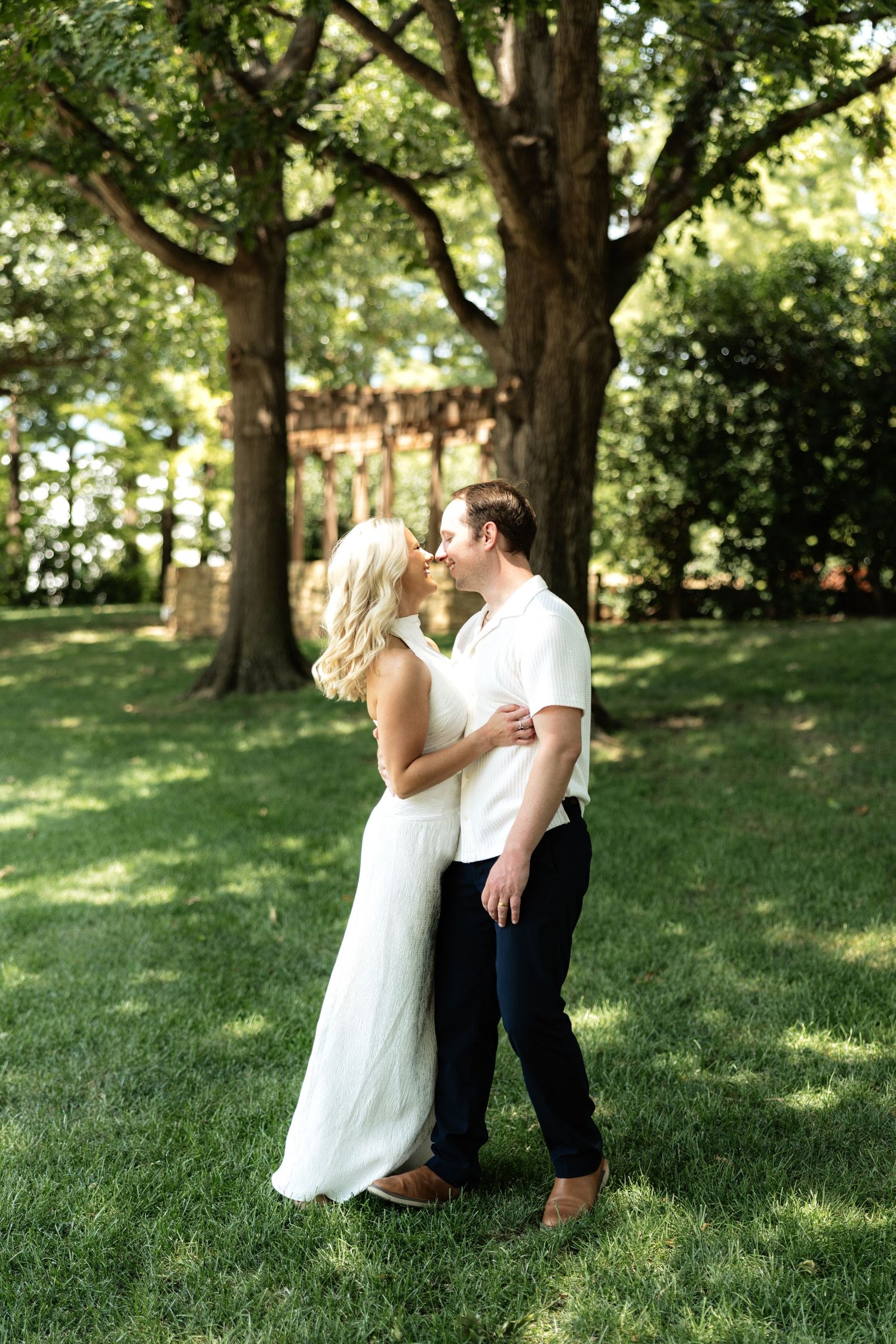 A couple shares a romantic kiss on a lush green lawn beneath large shade trees in an outdoor garden setting.