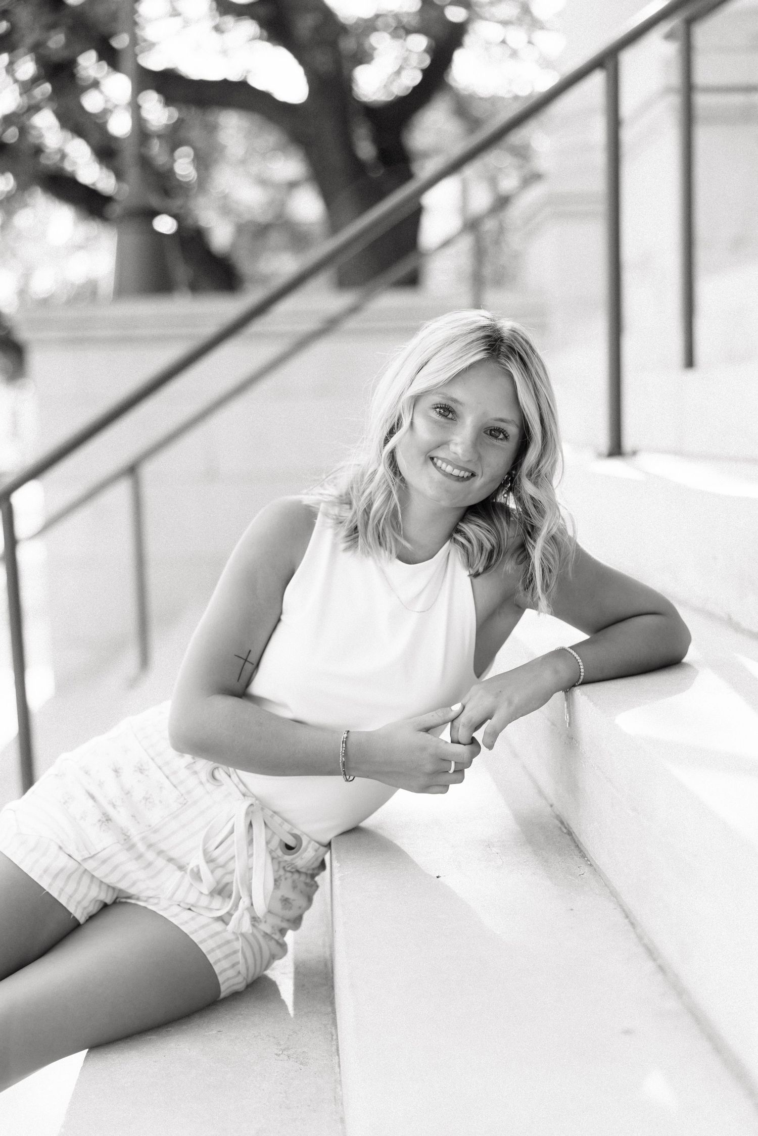 Black and white photo of a smiling blonde woman sitting on outdoor steps in a sleeveless top and shorts.