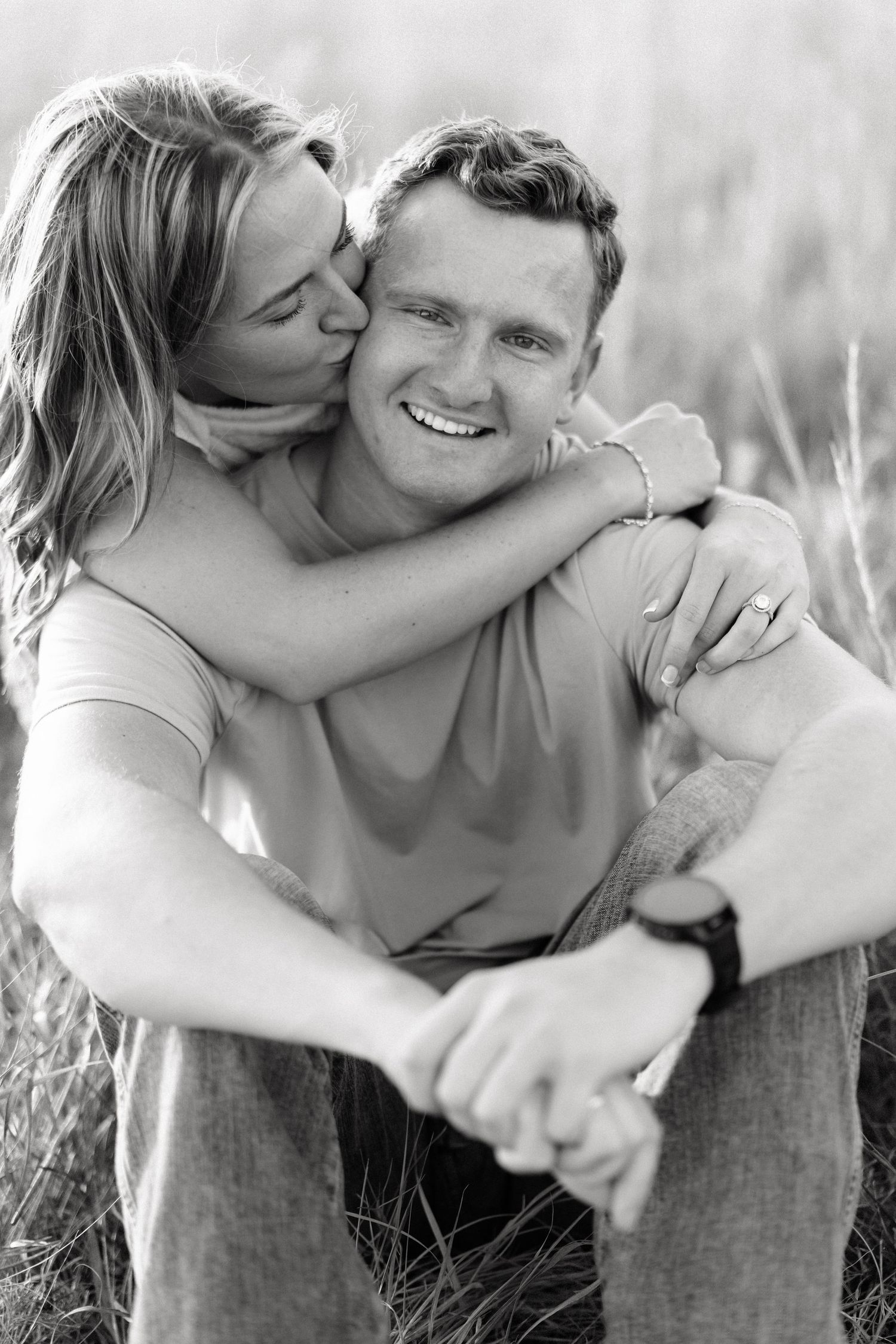 Black and white photo of a smiling man and woman embracing outdoors, she kisses his cheek while hugging him from behind.