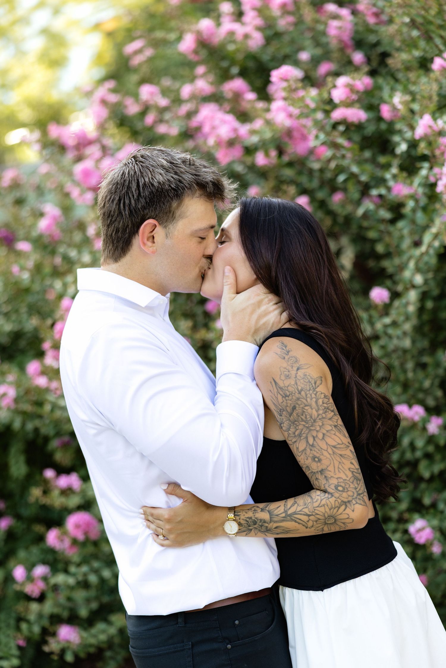 Couple kissing romantically in front of blooming pink roses, woman with tattooed arm in black top and white skirt.