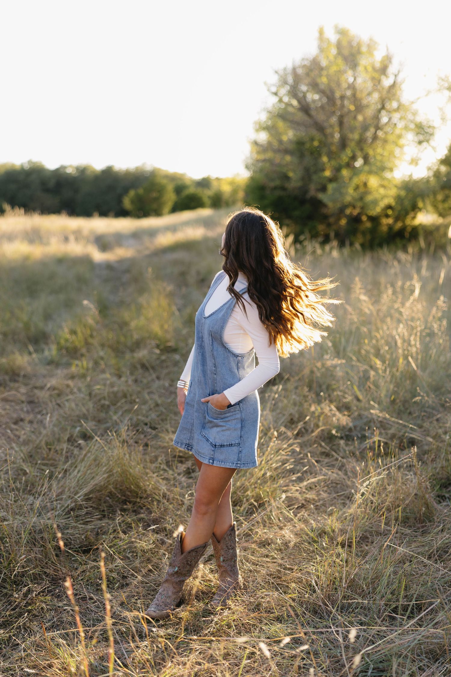 Brunette girl in denim pinafore and cowboy boots walking through golden field at sunset.