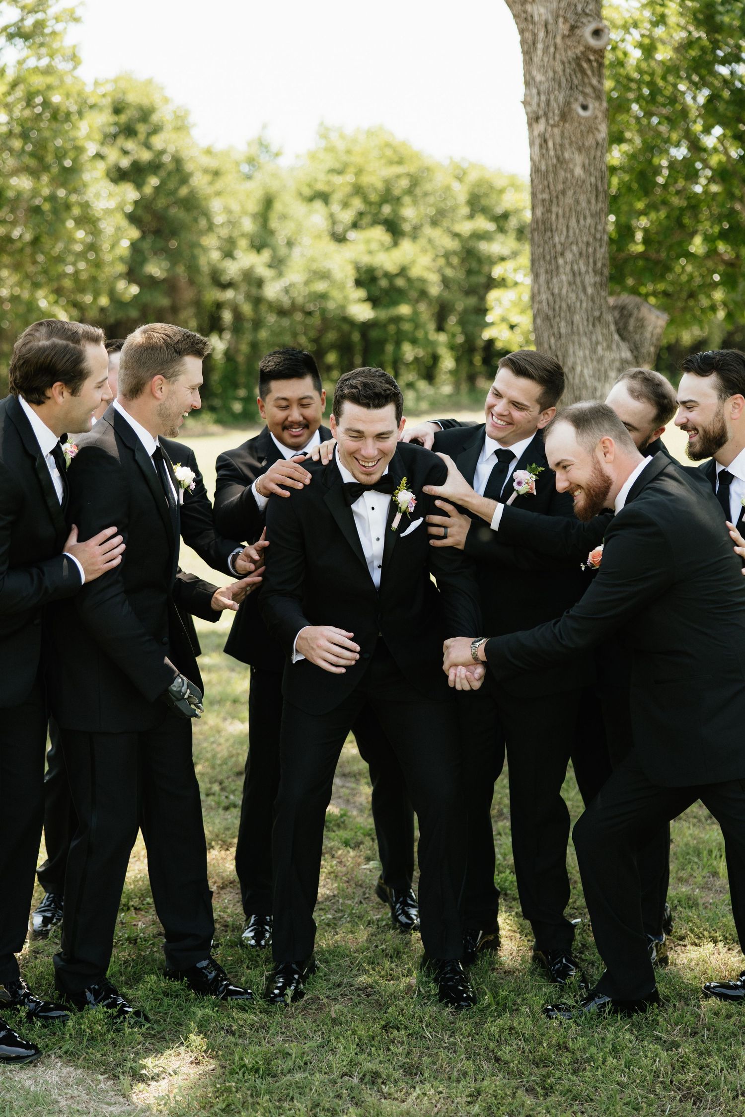 Groomsmen in black tuxedos laughing and celebrating with groom outdoors on wedding day.