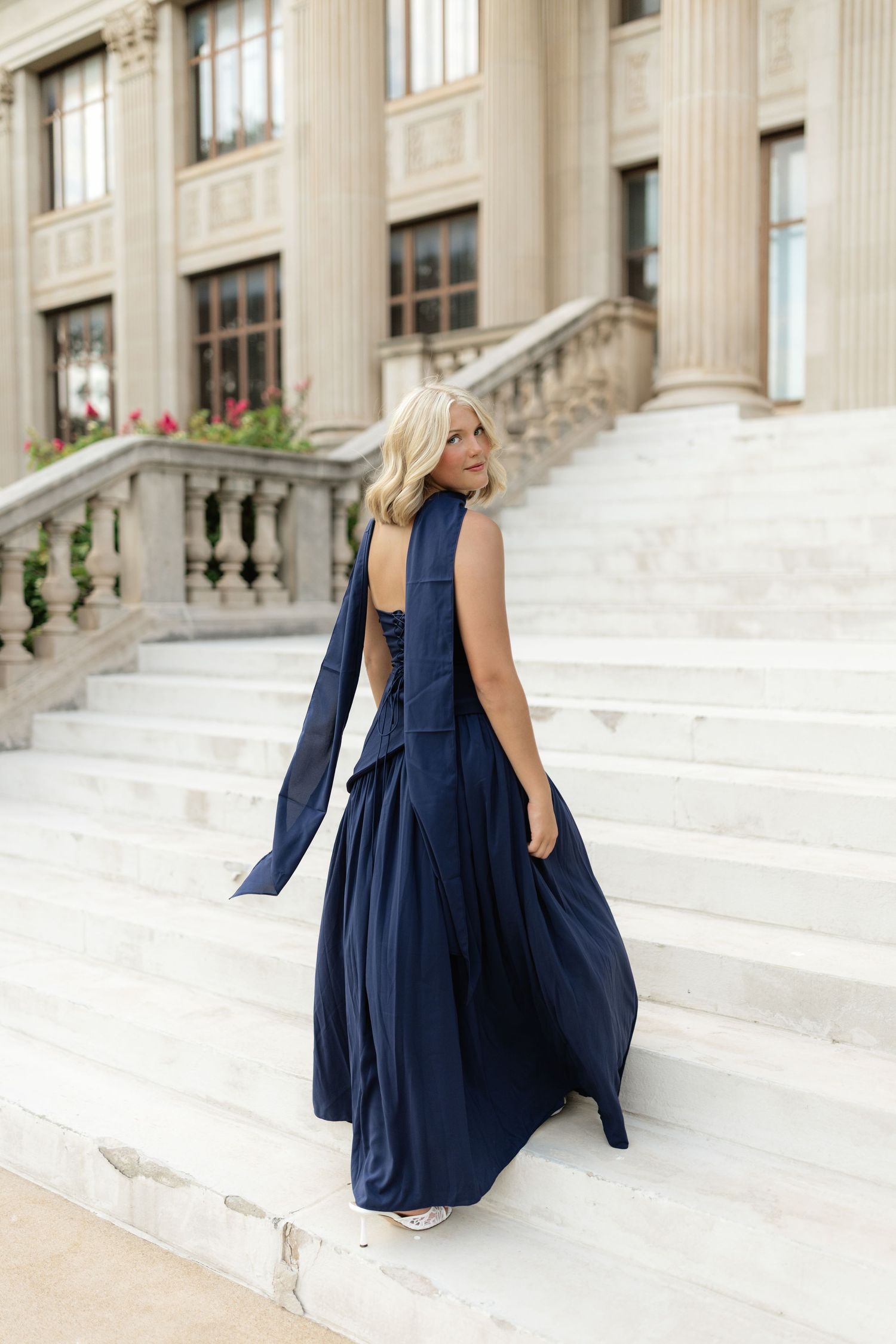 Woman in navy blue halter maxi gown poses on grand marble staircase of elegant building.