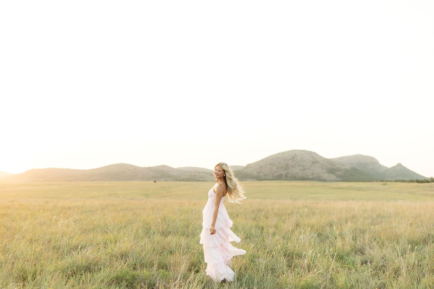 Woman in white flowing dress standing in golden sunlit field with mountain backdrop at sunset.