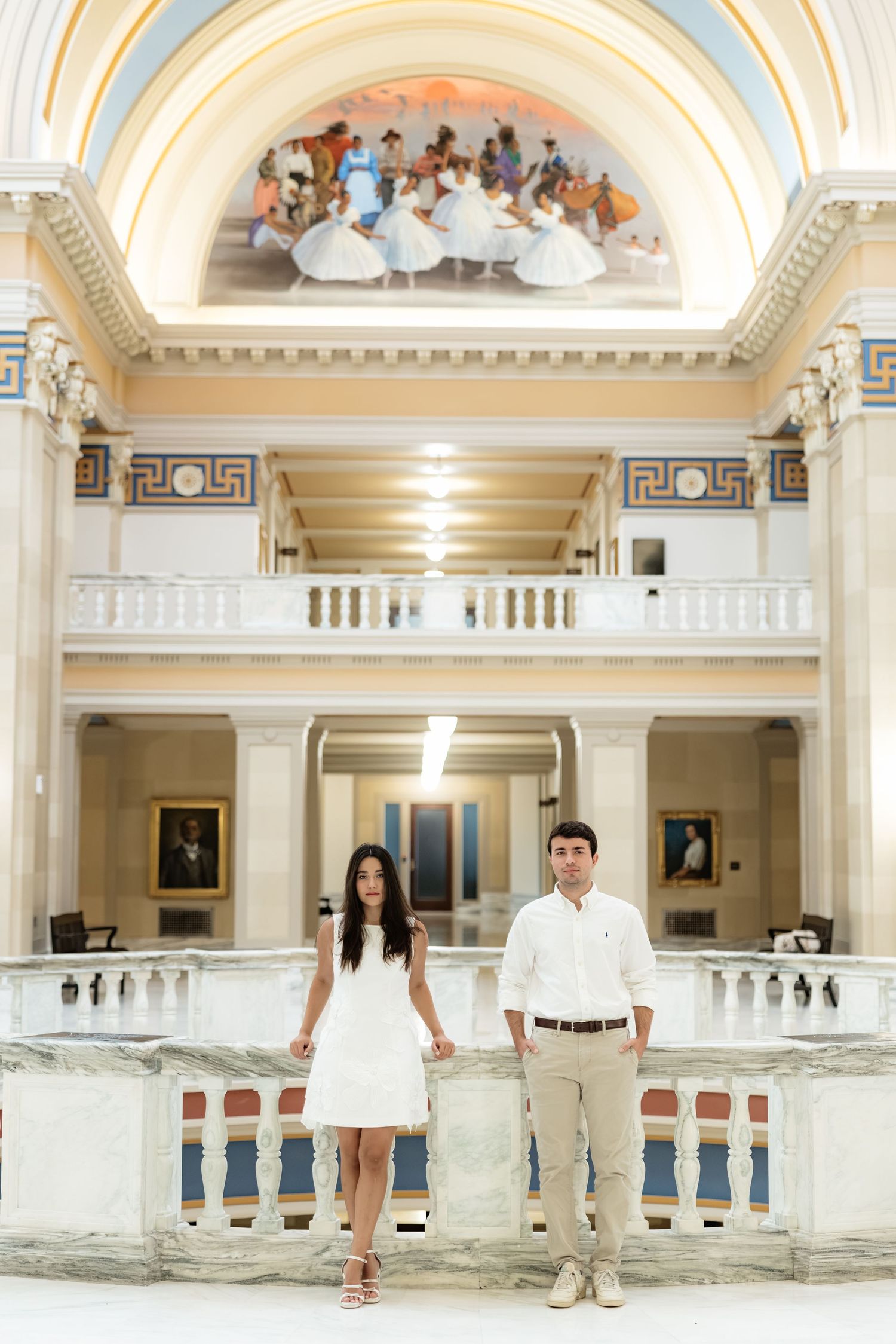 Couple in white attire posing in elegant neoclassical building with arched mural and marble balustrade.