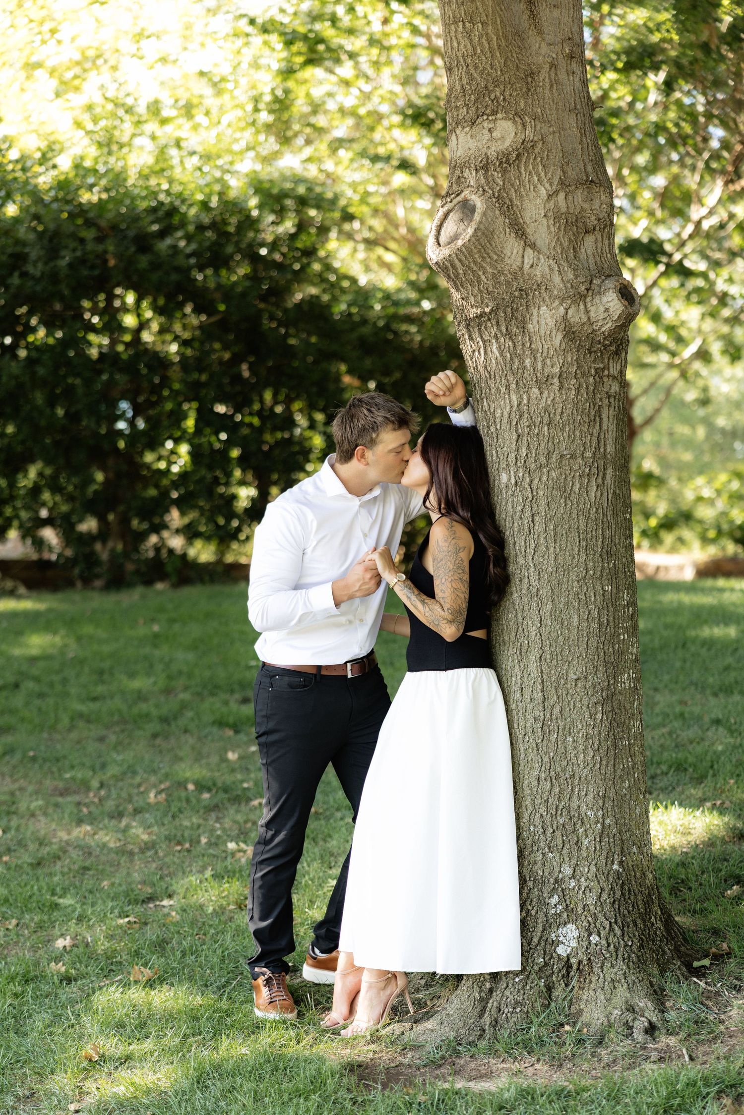 Romantic couple sharing a kiss by a tree in a lush green park, woman in white skirt and black top.