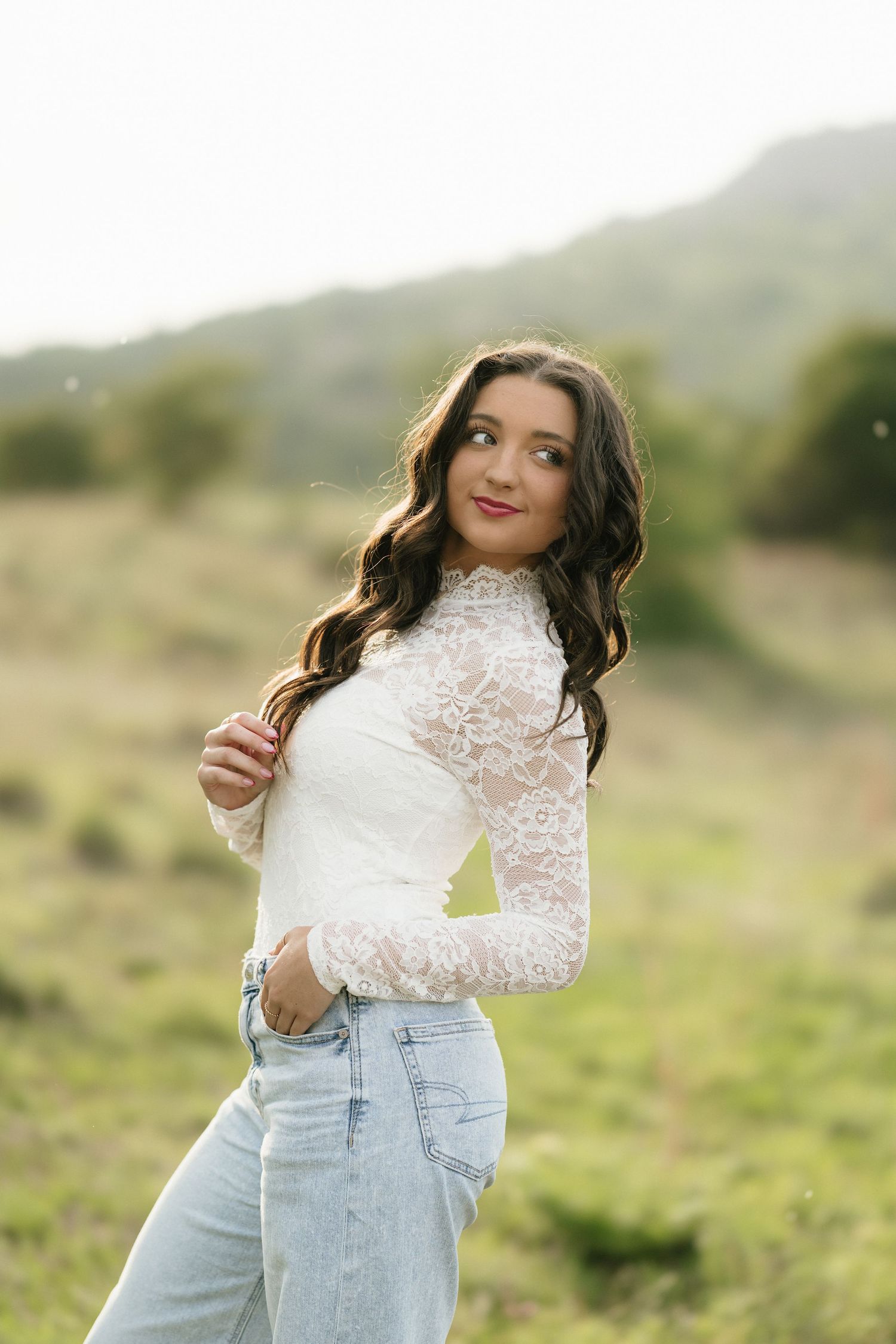 Brunette woman in white lace top and jeans poses gracefully in a scenic outdoor field with hills.