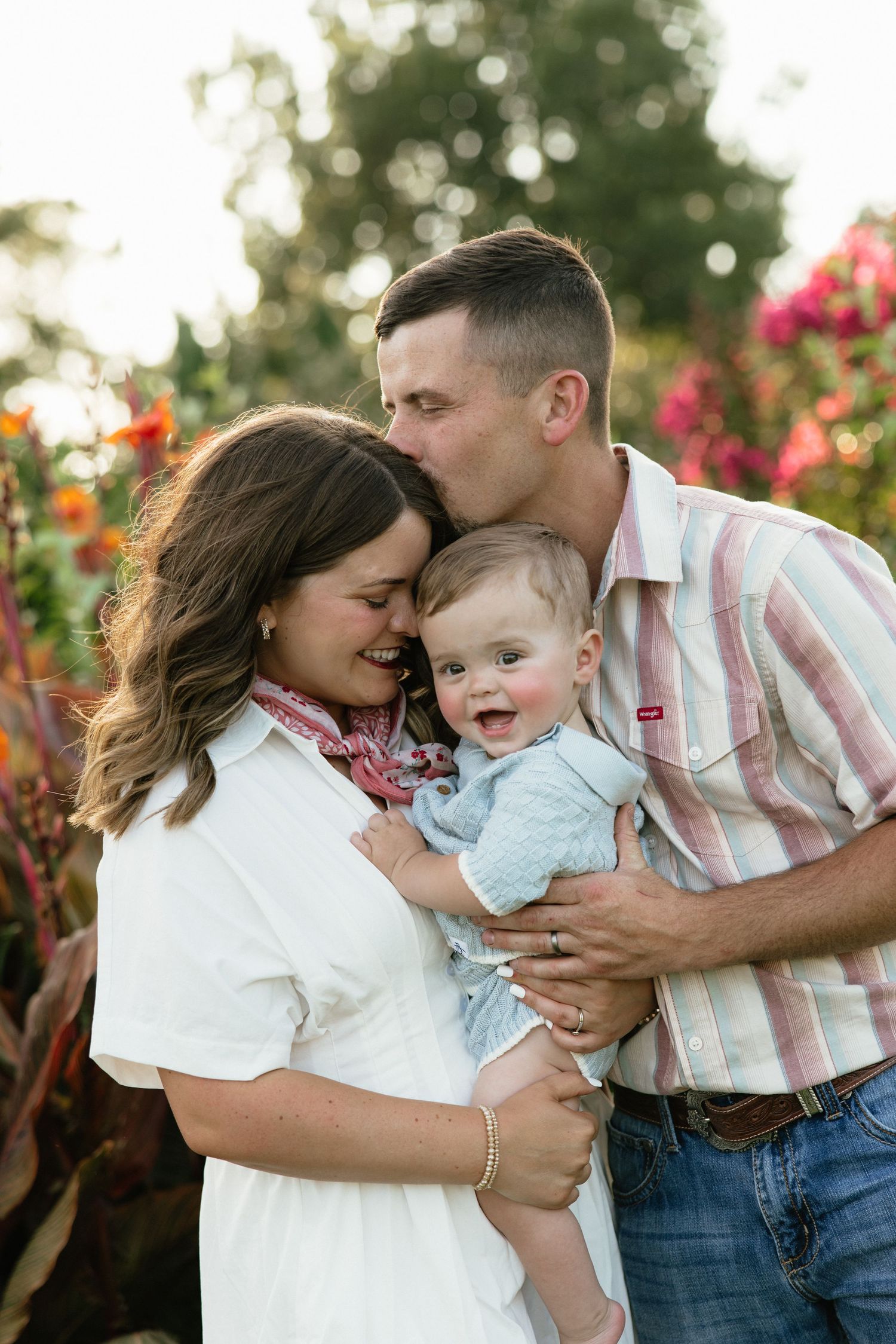 A happy family of three poses outdoors, with dad kissing mom while she holds their laughing baby boy.
