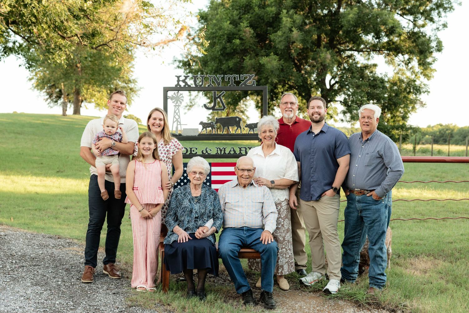 Multi-generational family poses outdoors near a farm sign, with elderly couple seated and others standing behind them.