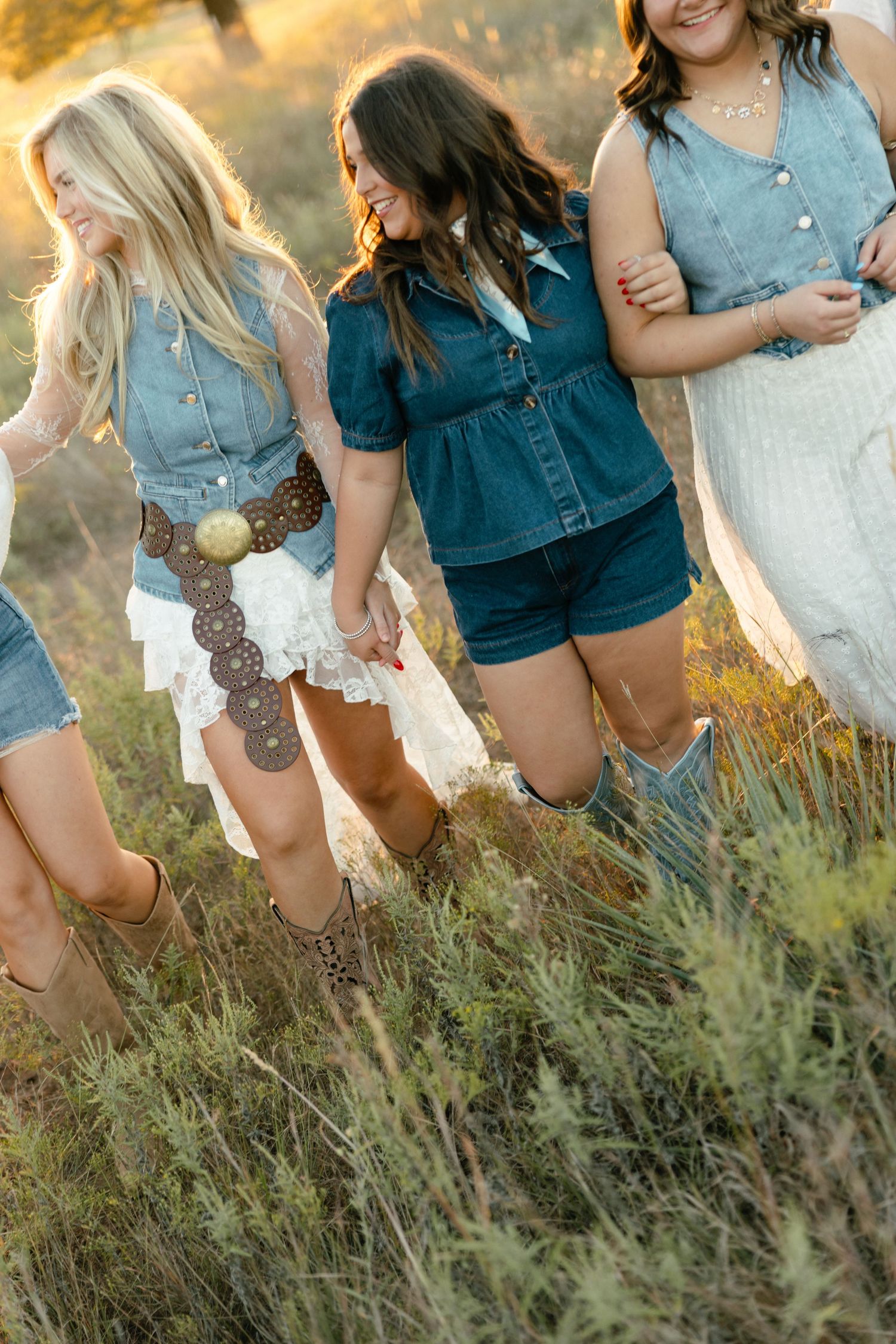 Three young women in western boho outfits walking together through tall grass at golden hour.