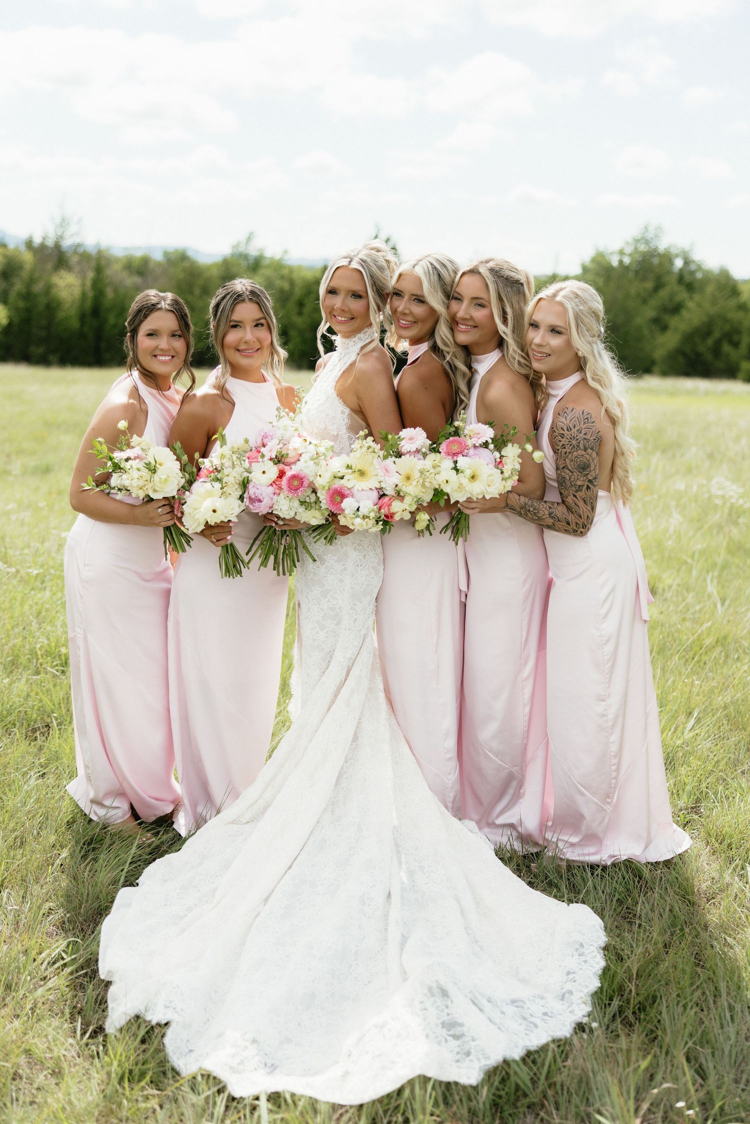 Bride in white lace gown with five bridesmaids in pink dresses holding floral bouquets in a green meadow.