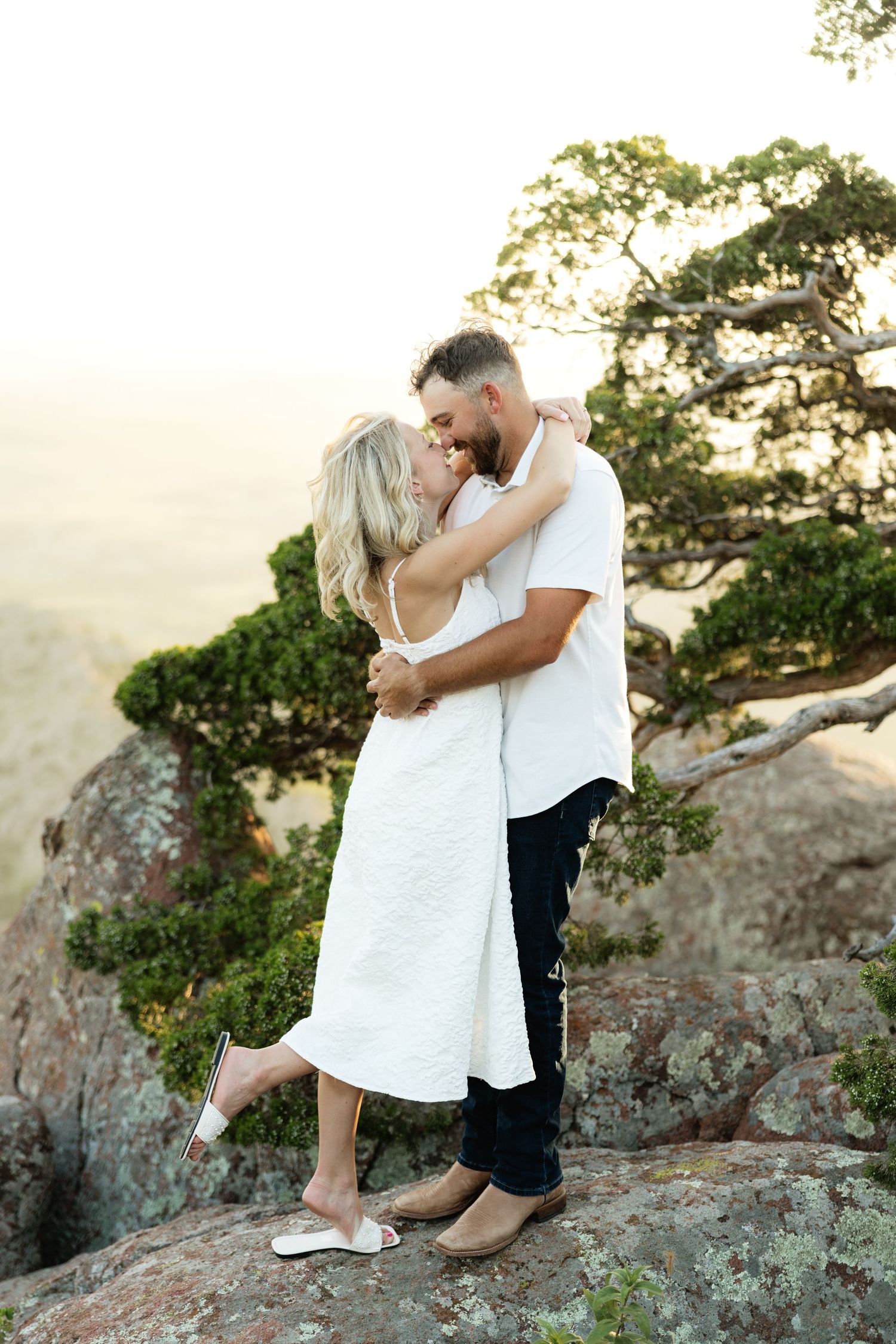 Couple embracing and kissing on a rocky mountain overlook with a scenic tree and misty valley backdrop.