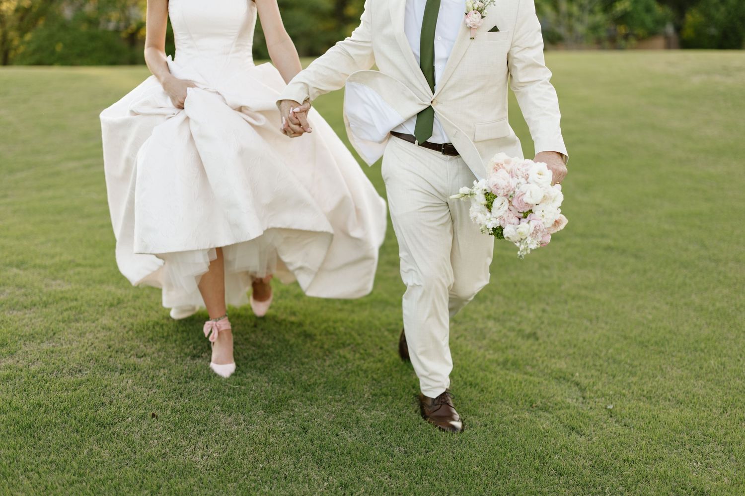 Bride and groom holding bouquet run hand in hand across a lush green lawn on wedding day.