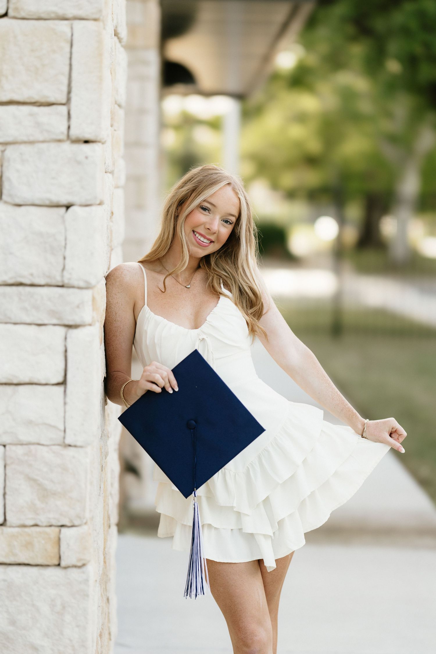 Smiling blonde senior poses with navy graduation cap in white ruffle dress outdoors.