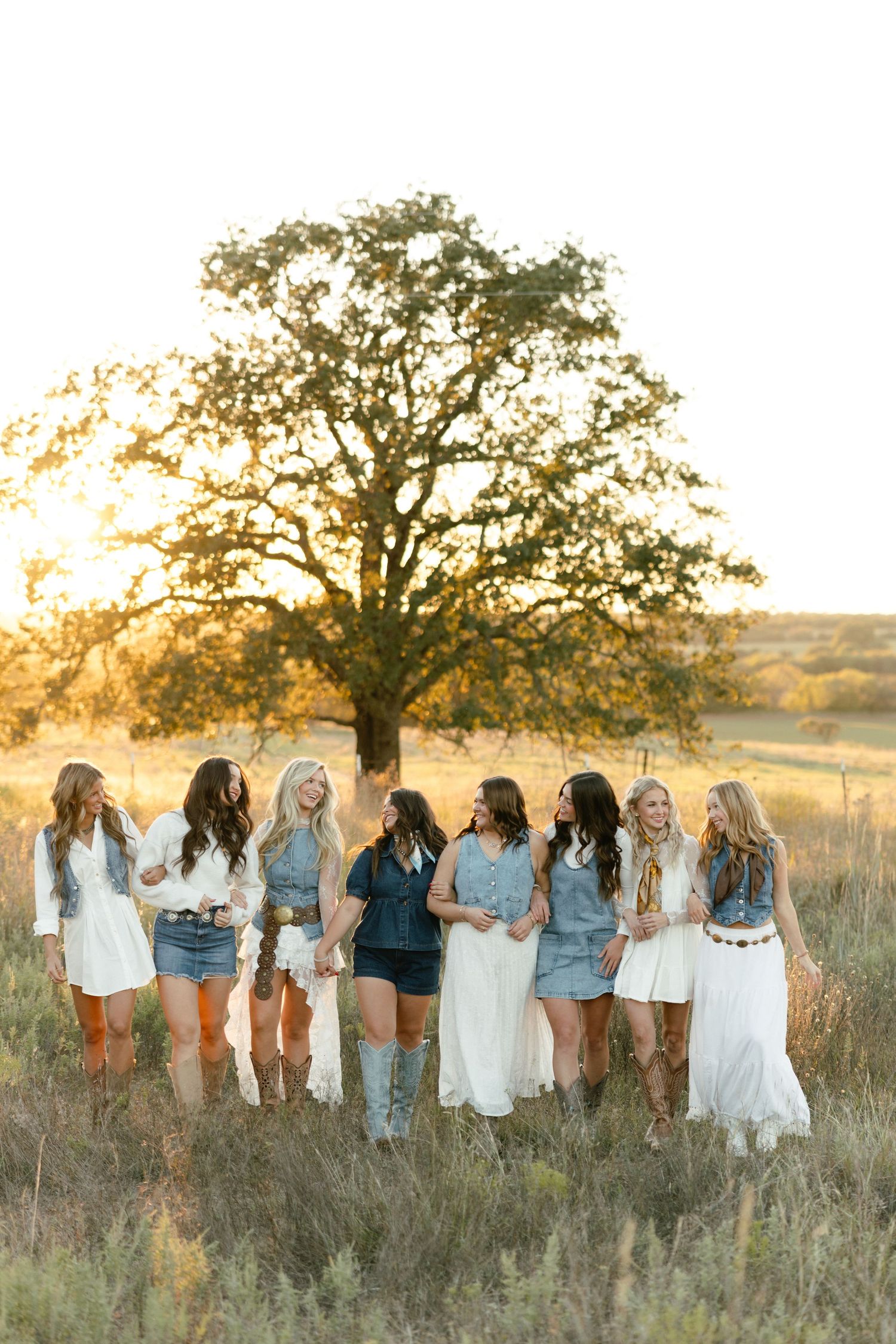 Bridal party in denim and white dresses with cowboy boots gathered under a large oak tree at sunset.