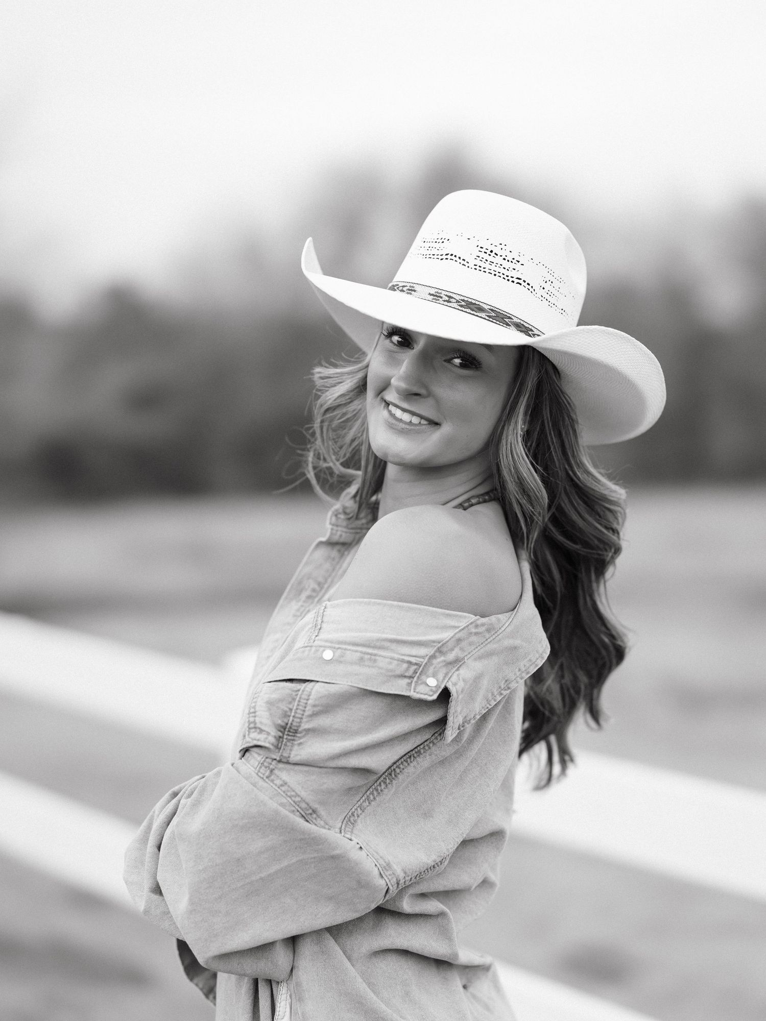 Young woman in white cowboy hat and denim top smiling over shoulder in black and white portrait.