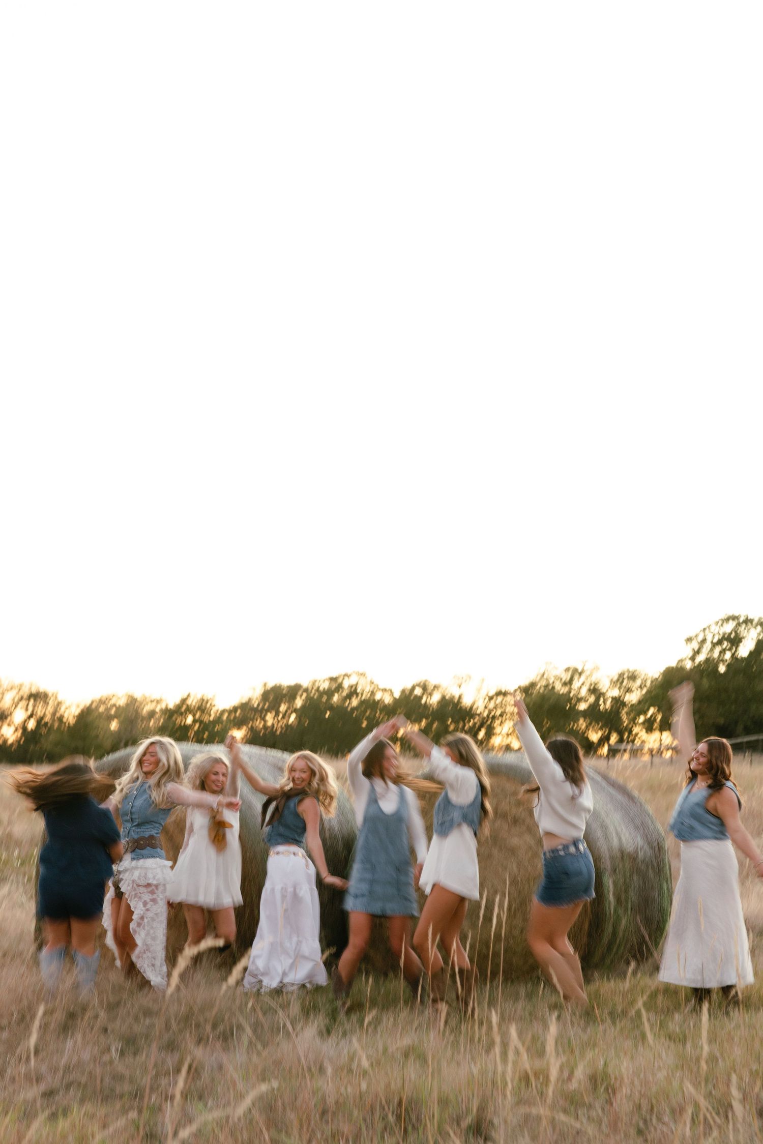 Group of young women dancing joyfully in a golden field at sunset near a hay bale.
