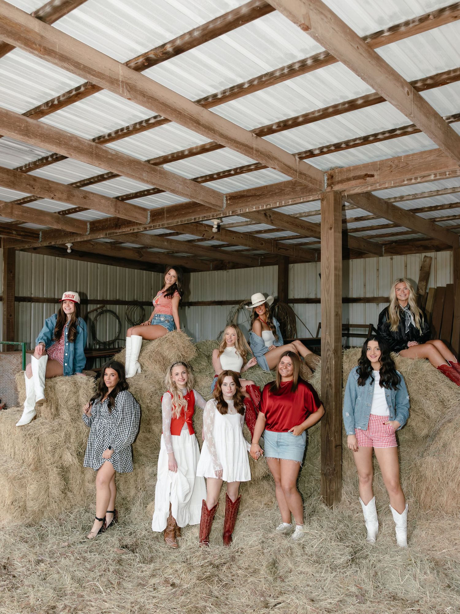 Group of women in western outfits posing together among hay bales inside a rustic barn.
