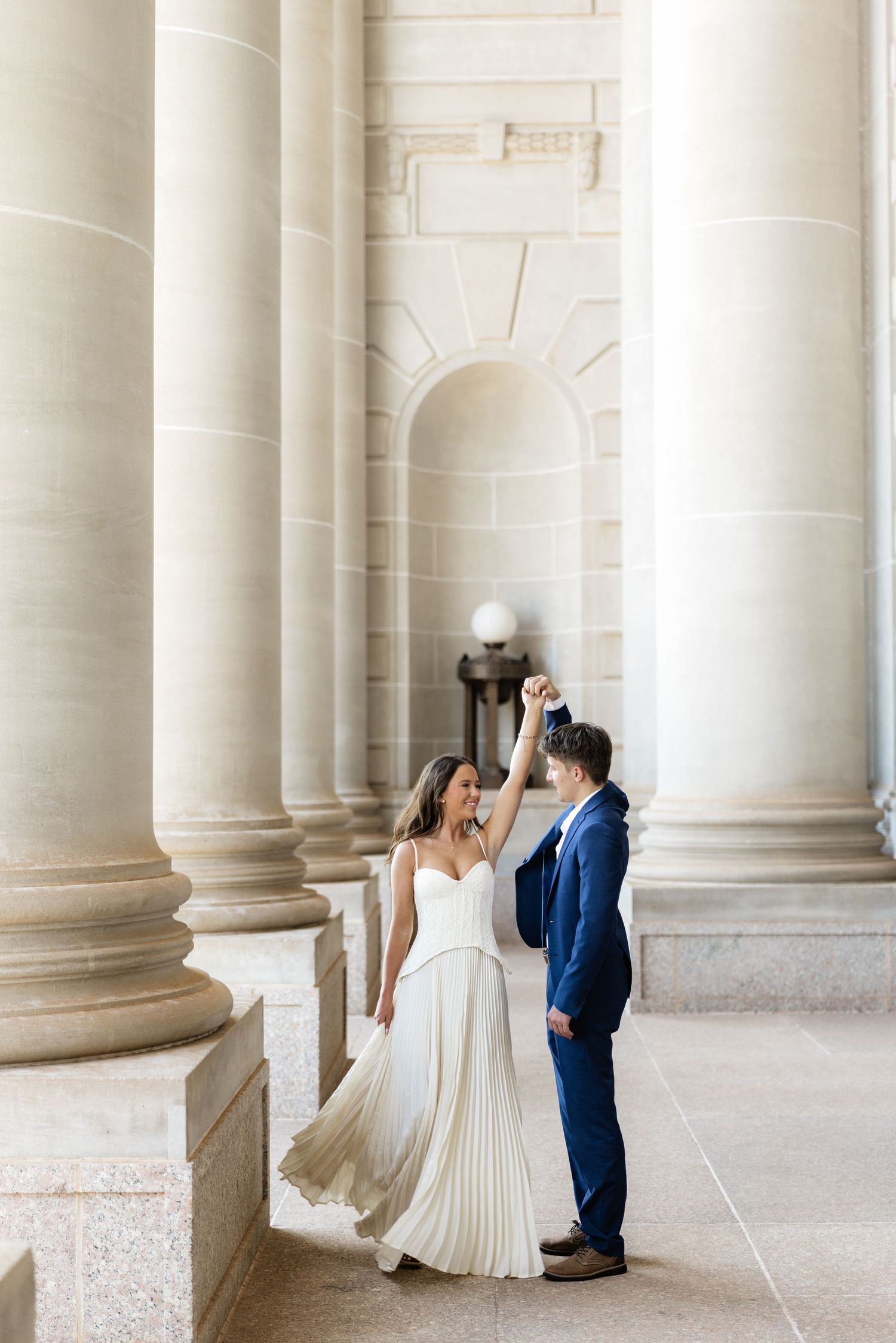 Bride and groom dancing among grand stone columns in elegant wedding attire, white pleated gown and navy suit.