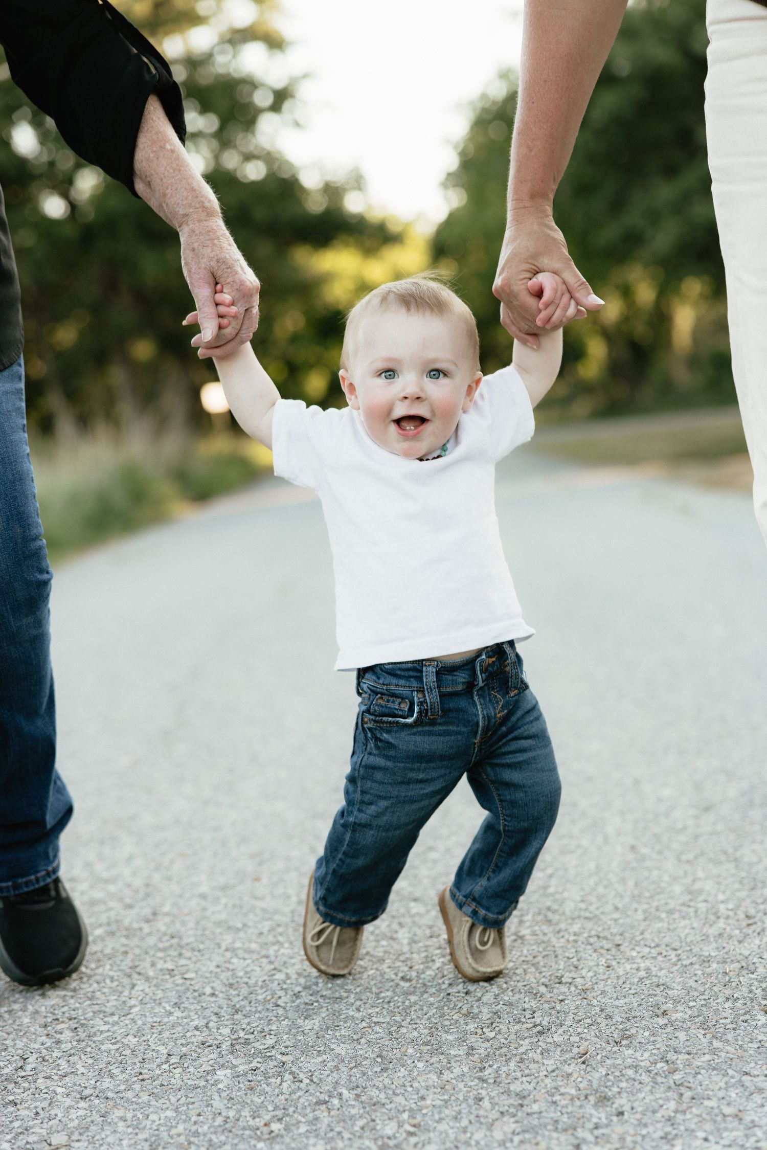 Happy baby boy in white shirt and jeans walks outdoors holding hands with two adults on a sunny path.